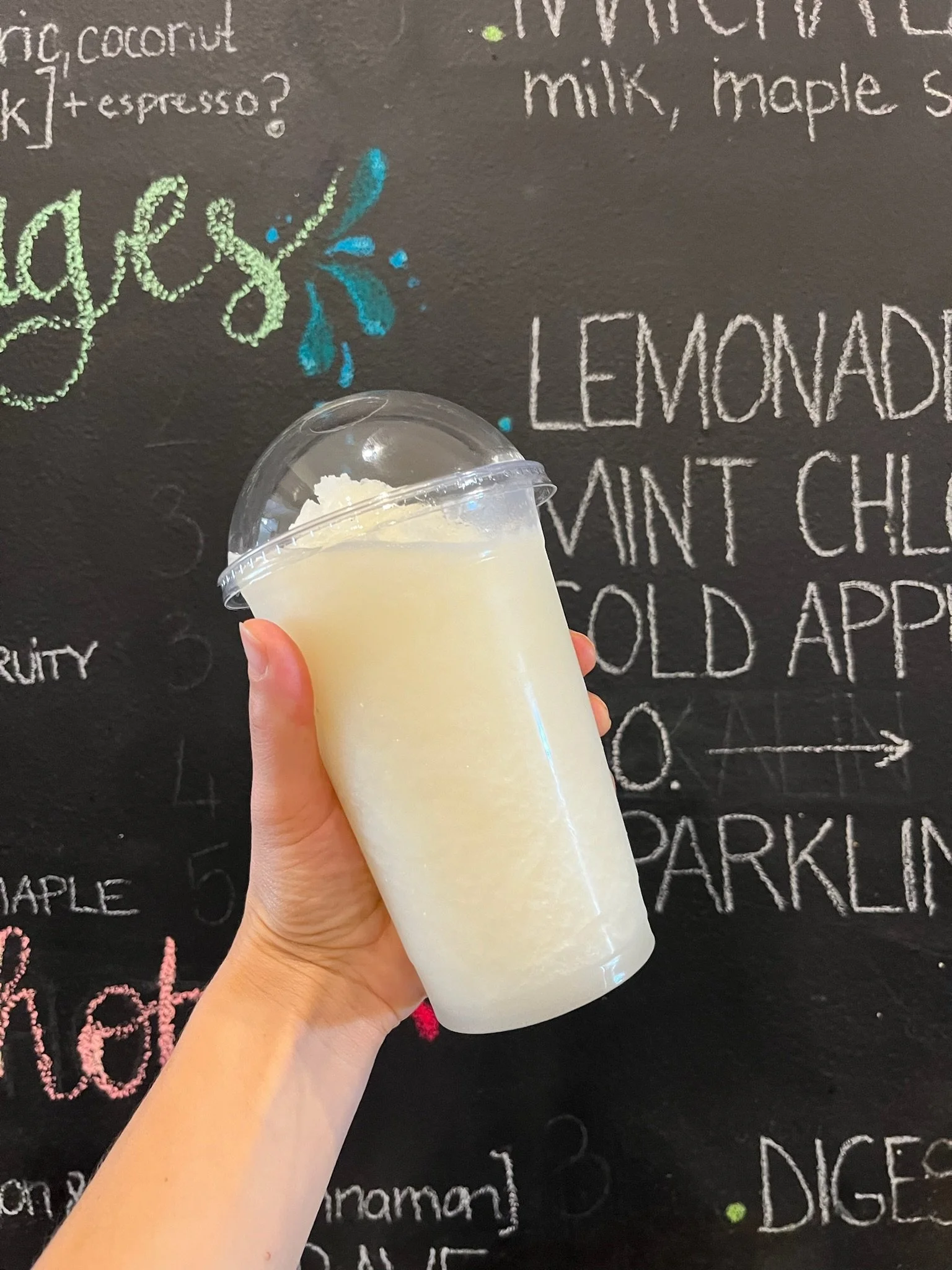 A hand holding a clear plastic cup filled with a white, frothy beverage, in front of a black chalkboard menu listing lemonades, cold drinks, and other beverages.