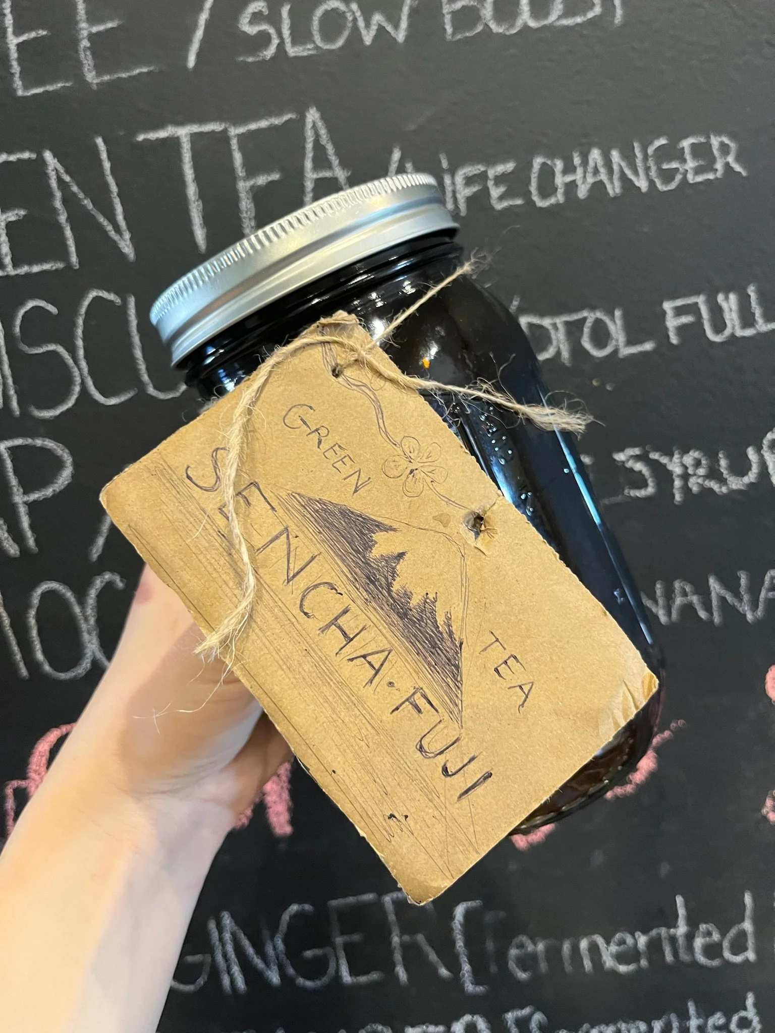 A dark glass jar with a metallic lid, wrapped with twine, and a brown hang tag that features an illustration of a mountain and the words 'Green Tea' and 'Fujii.' In the background, a chalkboard with handwritten words and phrases.