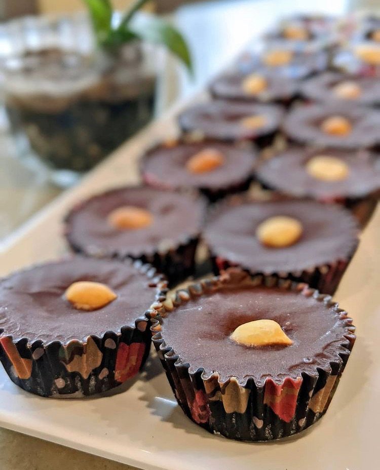 Chocolate cups topped with a single almond on a white platter, with a blurred background including a potted plant.