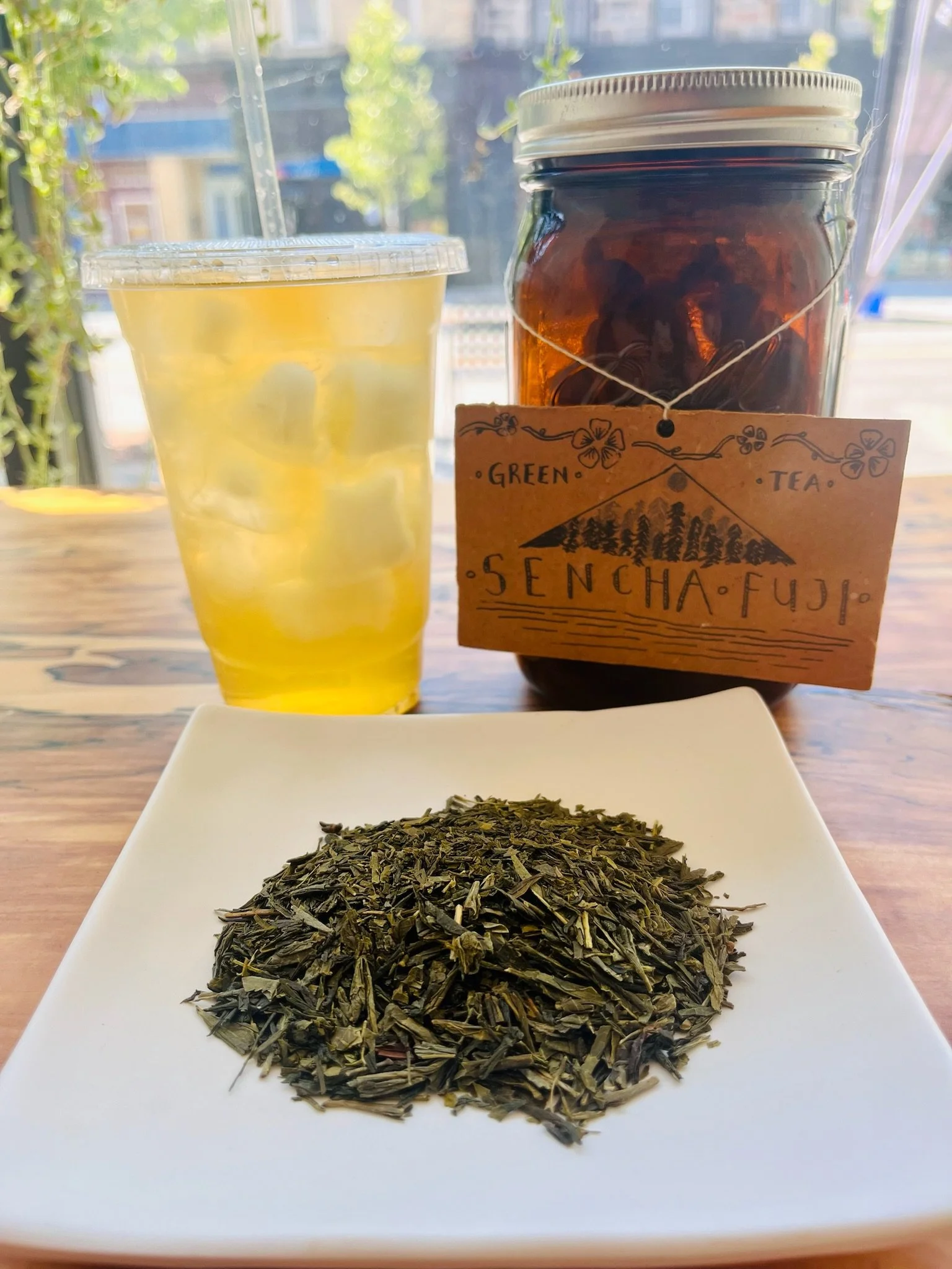 A plate of loose green tea leaves on a white square dish, a glass of iced tea with lemon slices, and a jar of brewed tea labeled 'Sencha Fuji' with a handwritten tag, set on a wooden table in front of a window with sunlight.
