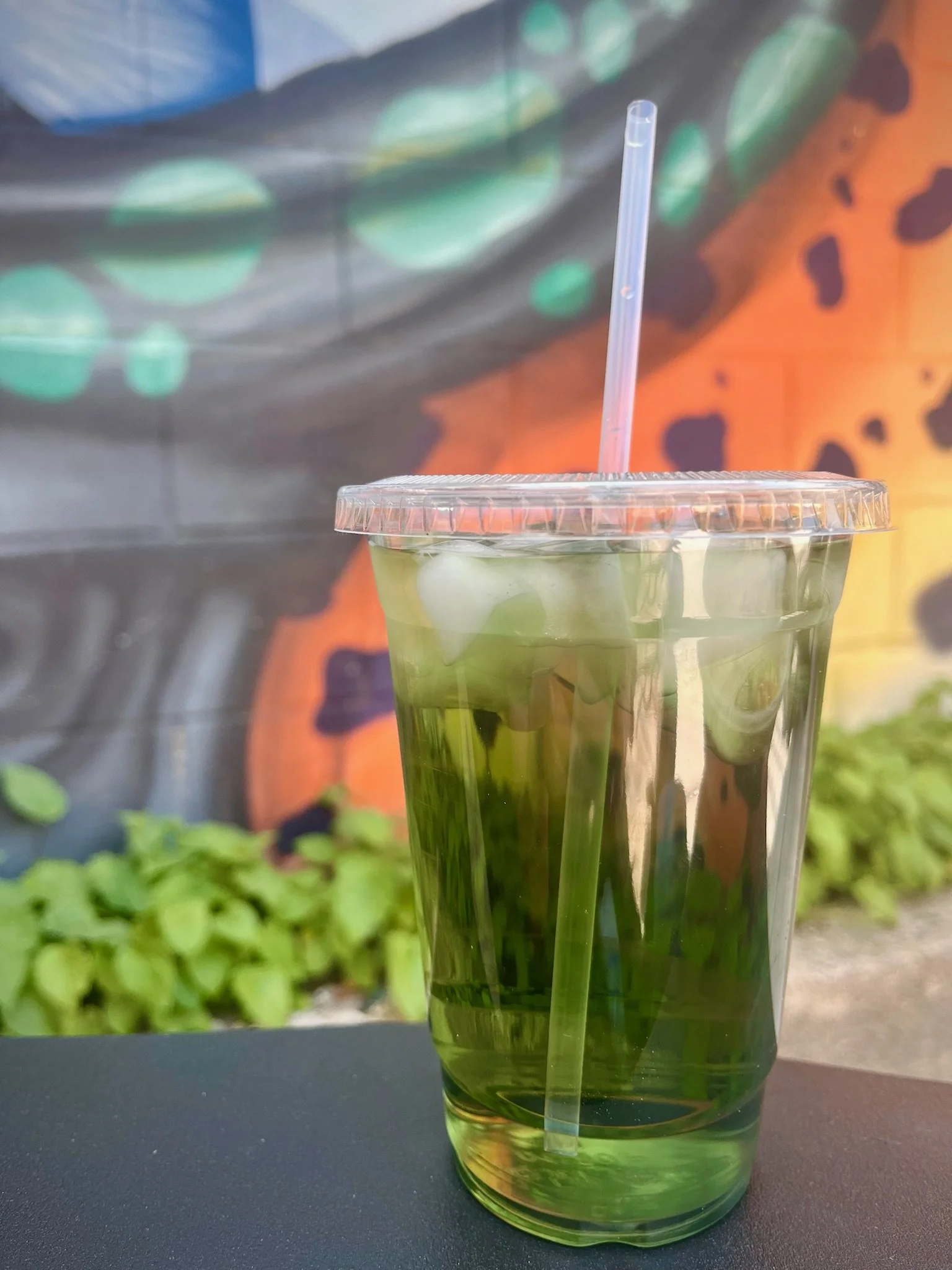 Close-up of a plastic cup filled with green liquid and a straw, set against a colorful graffiti wall background.