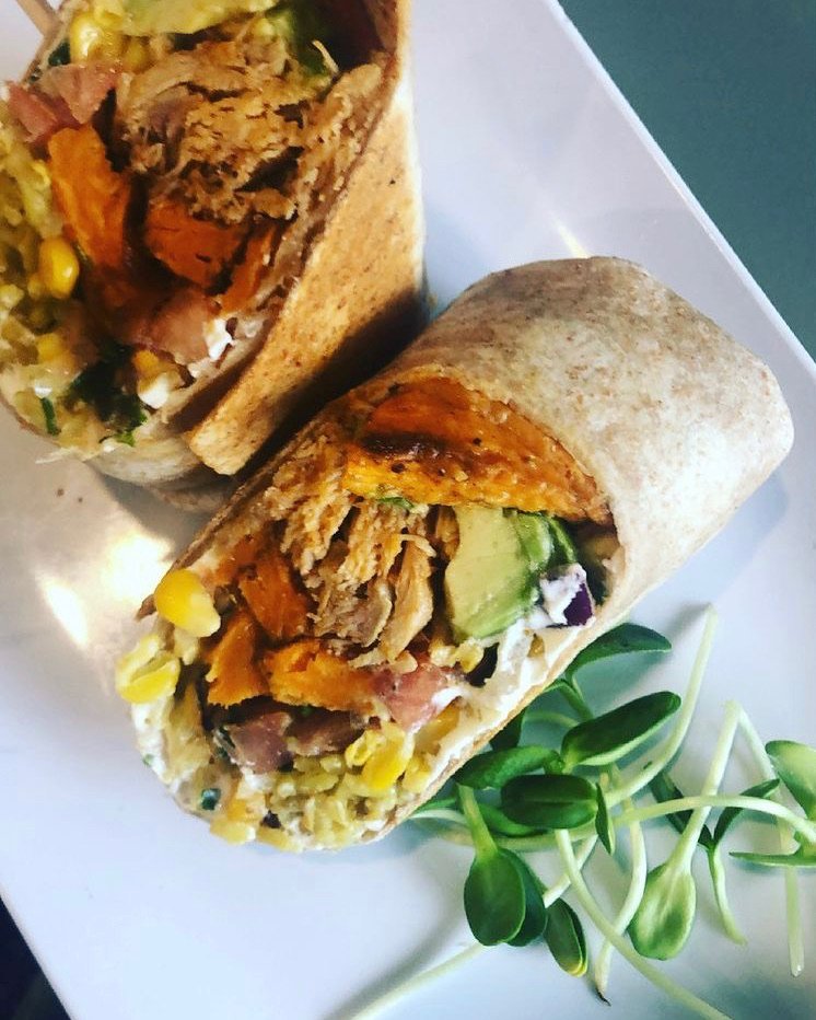 A sliced wrap sandwich on a white plate, filled with vegetables, meat, corn, and sauce, with some microgreens on the side.
