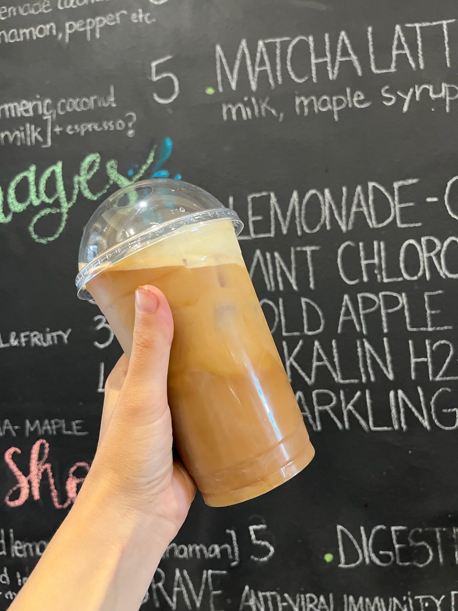 A person's hand holding a plastic cup filled with iced matcha latte against a black chalkboard menu background.