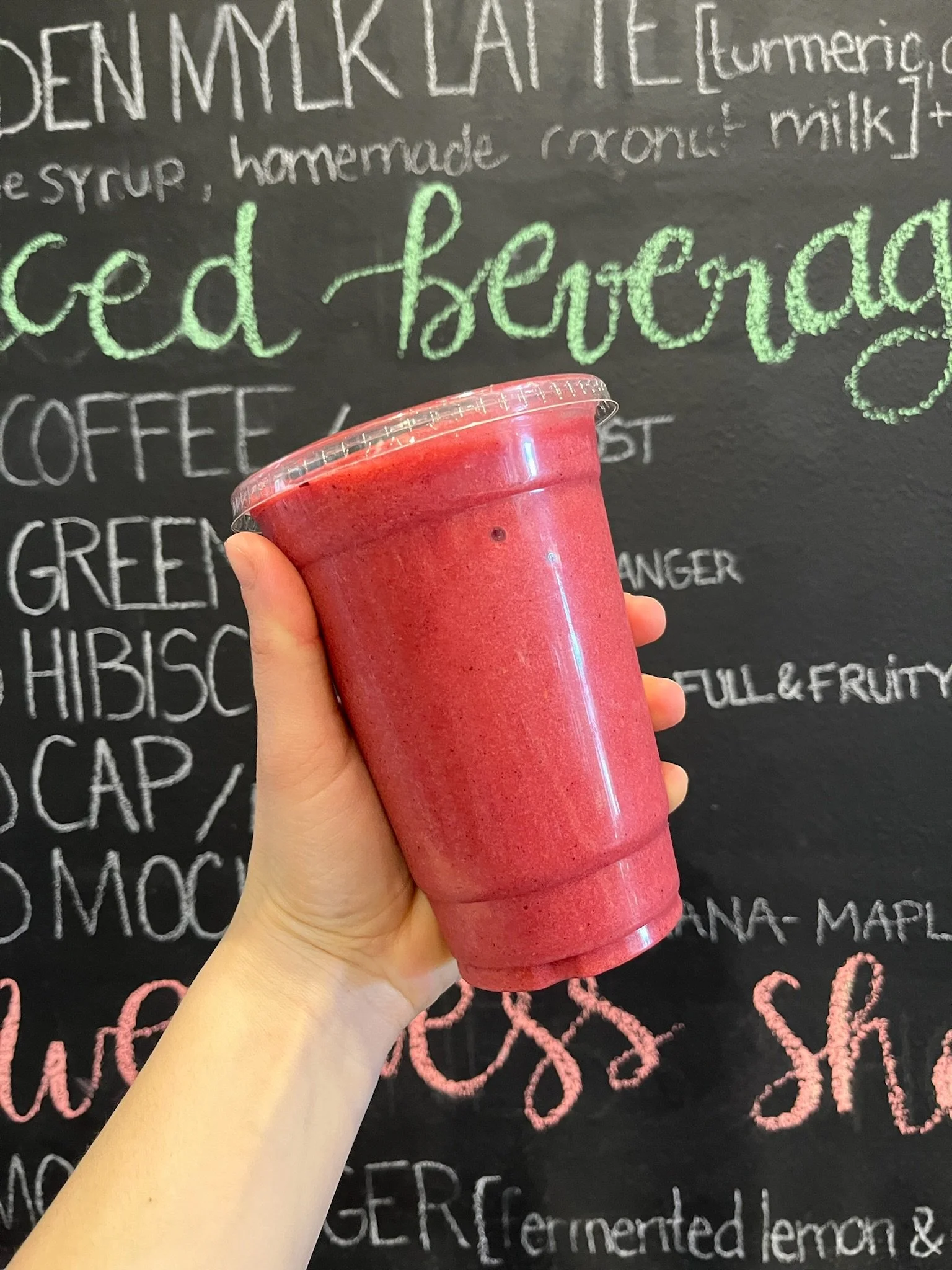 A hand holding a red smoothie in a clear plastic cup with a domed lid in front of a black chalkboard menu with colorful chalk writing of various flavors.