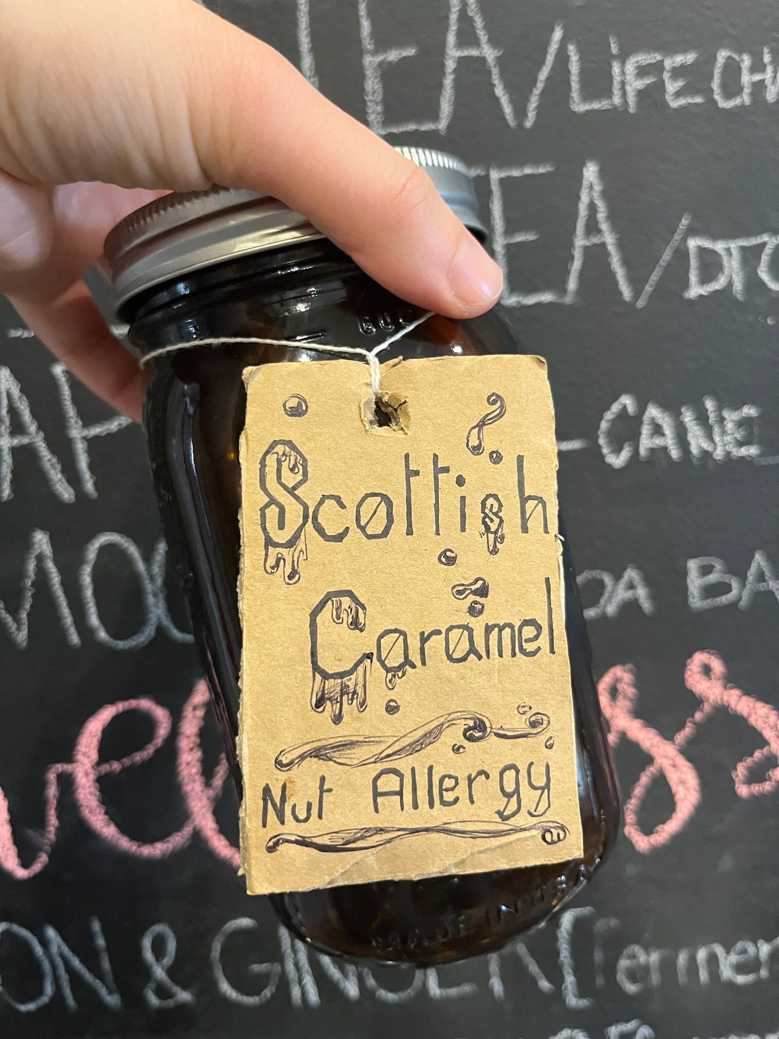 A small dark glass jar with a metal lid, labeled 'Scottish Caramel' with a handwritten tag indicating it is nut allergy safe, hanging from a string.