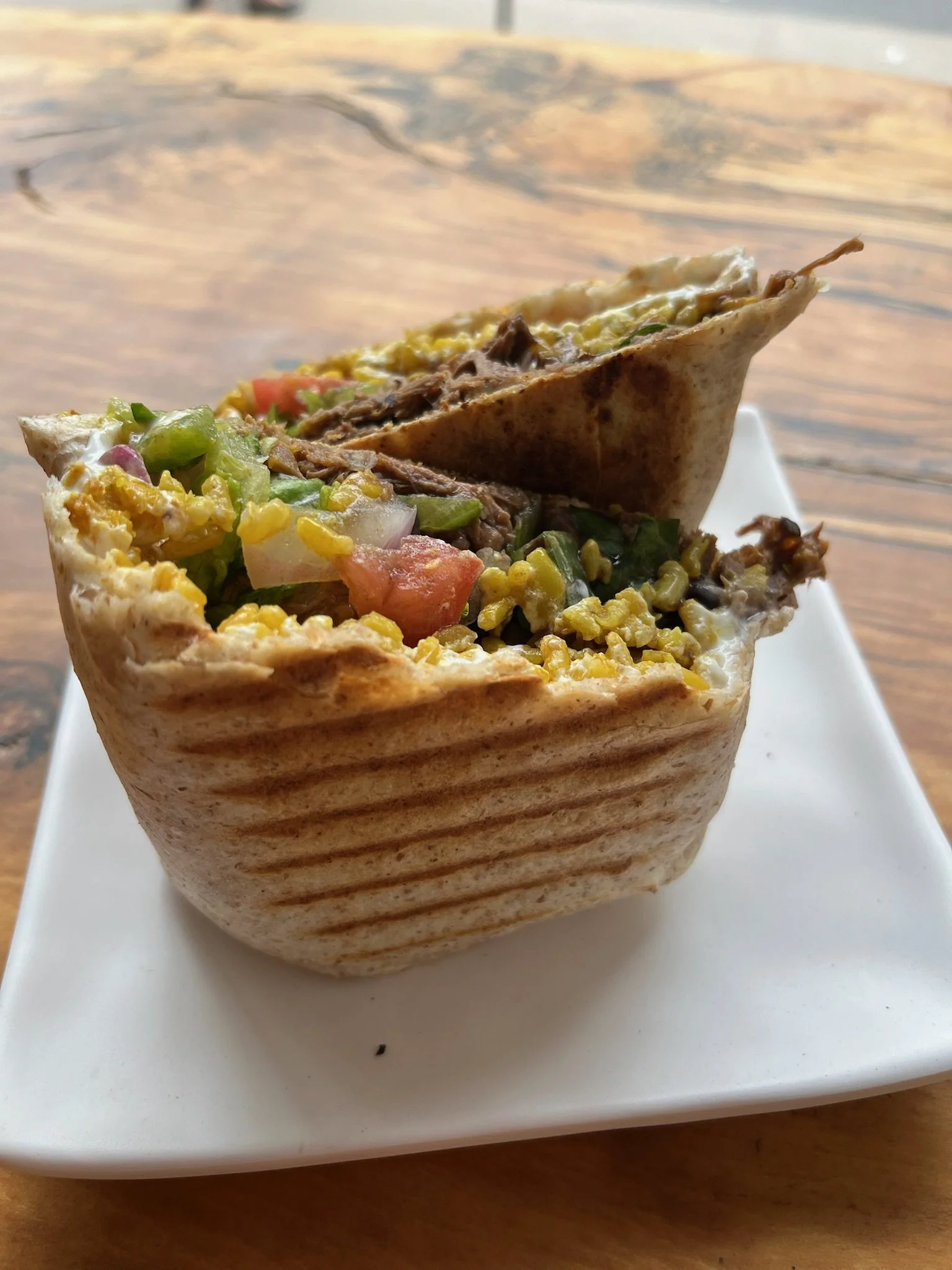 A close-up of a grilled wrap with visible grilled marks, filled with shredded beef, chopped vegetables, corn, and melted cheese, served on a white square plate on a wooden table.