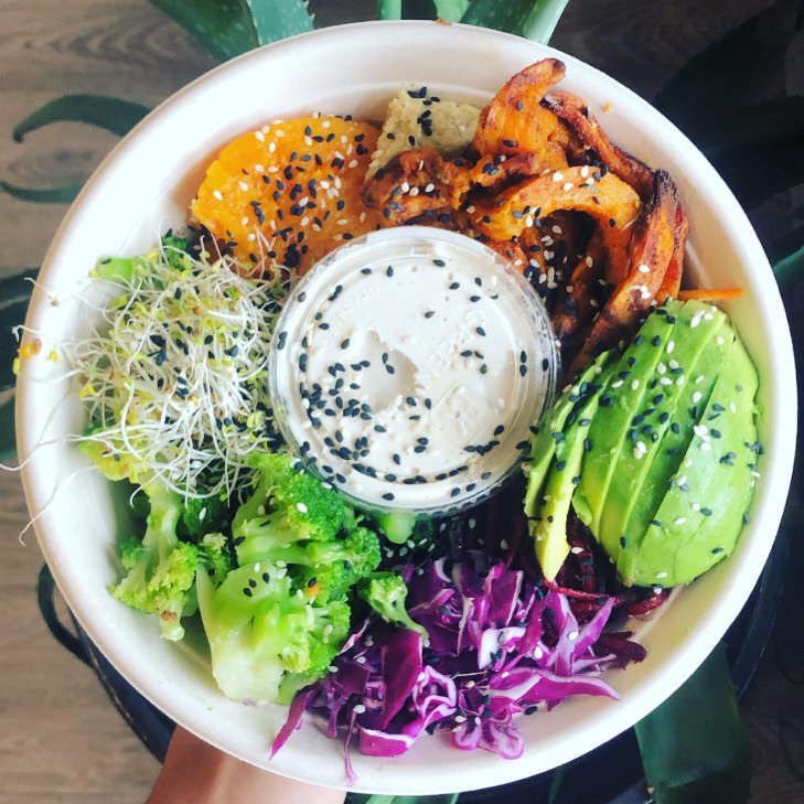 A colorful salad bowl with mixed greens, purple cabbage, sliced avocado, grilled chicken, broccoli, shredded carrot, microgreens, a dollop of sauce with black sesame seeds, and other vegetables.