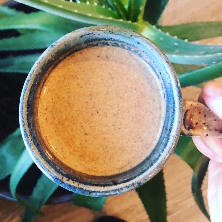 A ceramic mug of frothy coffee being held over a wooden table with green plant leaves in the background.