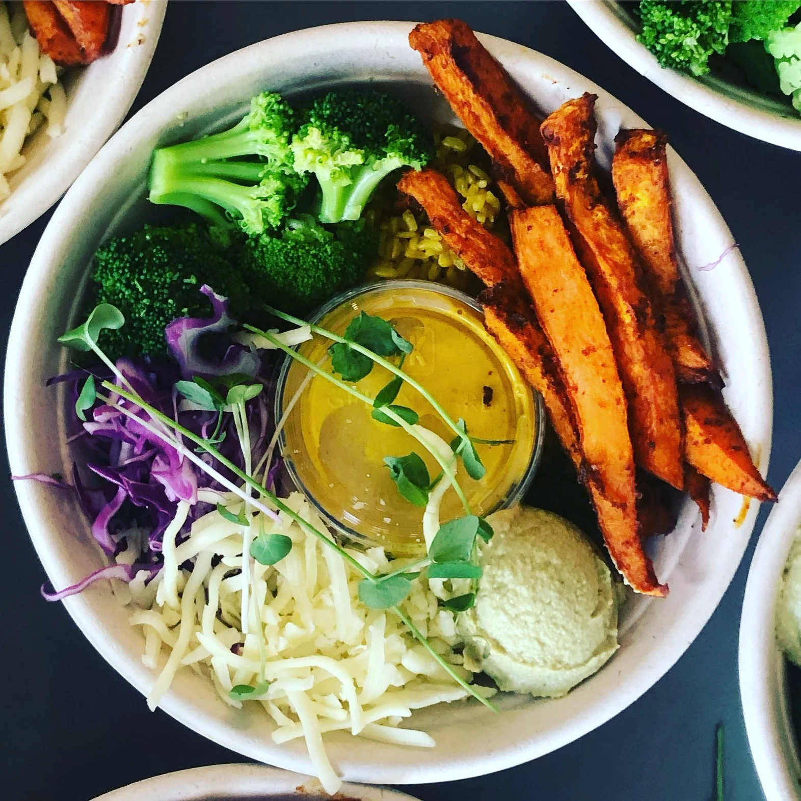 A bowl of roasted sweet potato fries, broccoli, purple and white shredded cabbage, shredded cheese, a dollop of hummus, microgreens, and a small container of olive oil with herbs.
