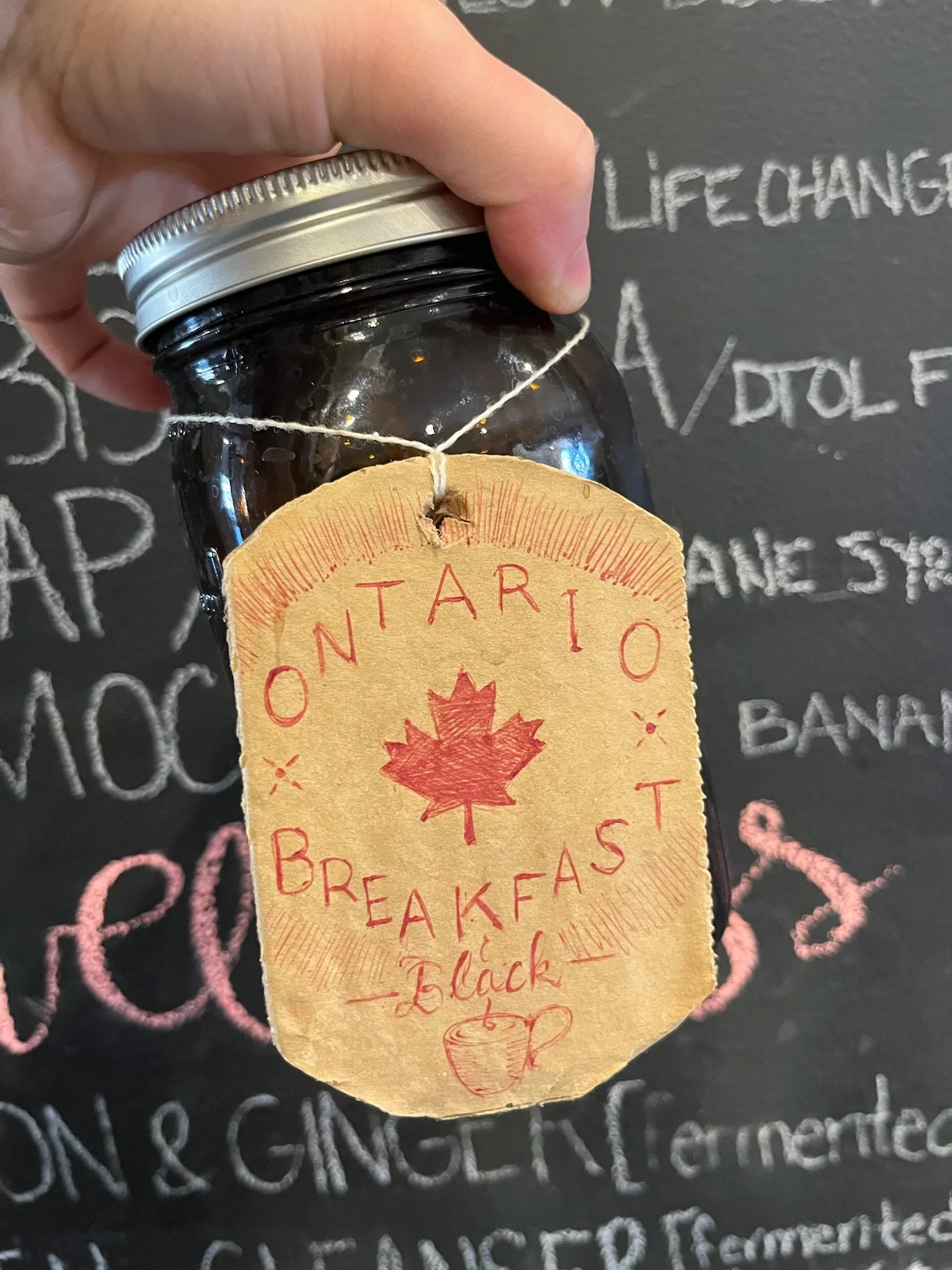 A glass jar with a metal lid, labeled 'Ontario Breakfast - Black,' hanging from a string, held by a person in front of a blackboard with handwritten chalk text.