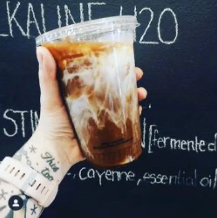 A hand holding a plastic cup of iced coffee with milk and swirling coffee sitting in front of a chalkboard with writing, including the word 'KALINE' and the number '20'.