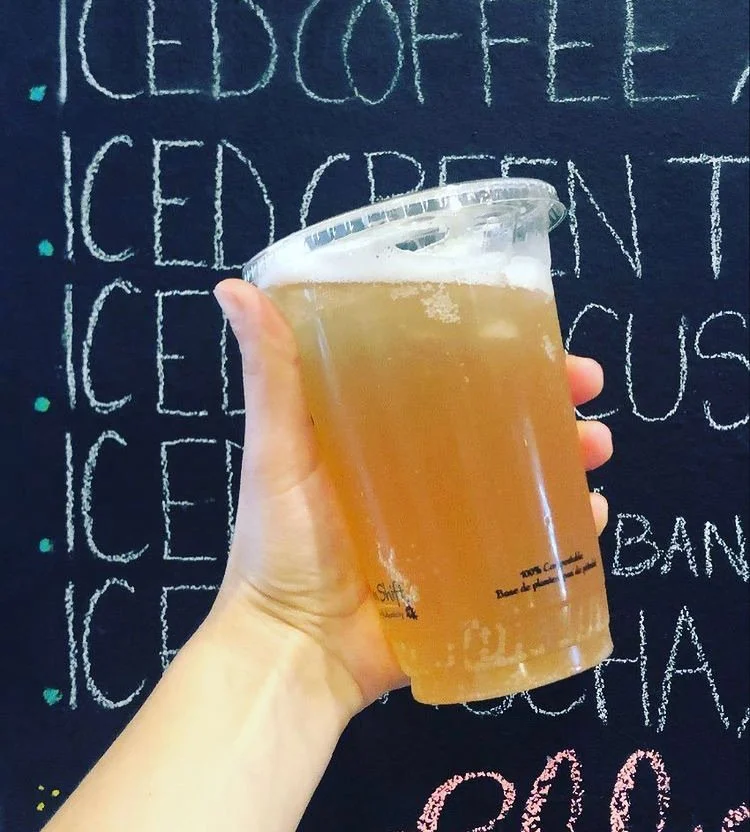 A person holding a clear plastic cup of beer in front of a chalkboard menu listing various iced beverages.
