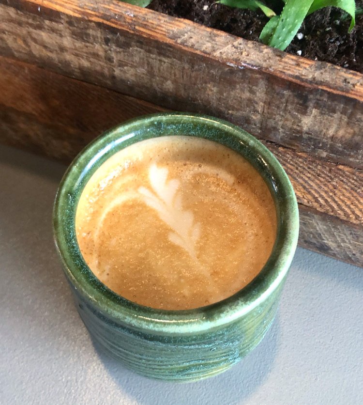 A green ceramic mug with a latte featuring latte art in the shape of a leaf, placed on a gray surface next to a wooden box containing plants.