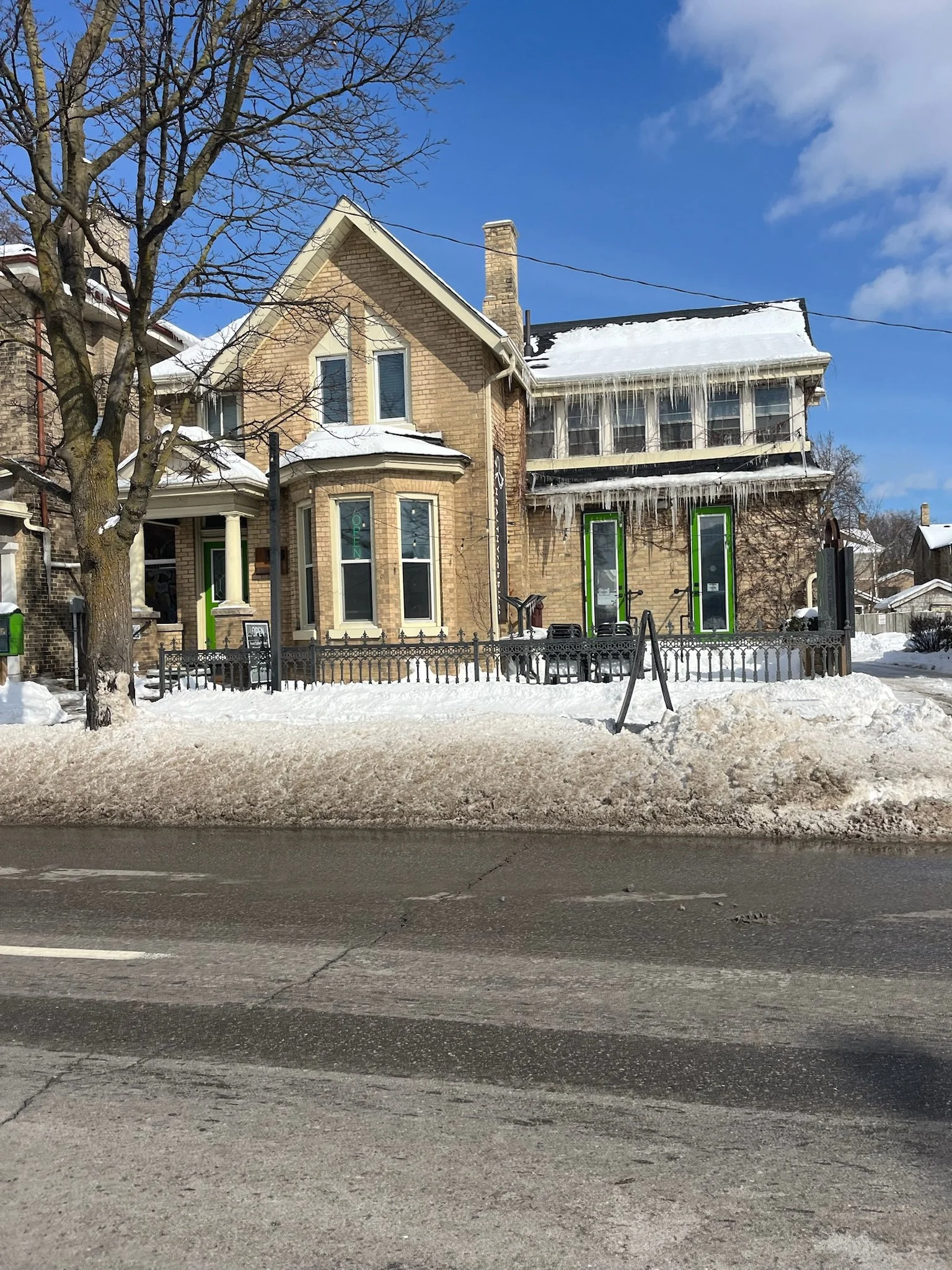 A snowy street view of two-story brick houses with icicles hanging from the roof. Two houses have large windows and a small front porch, with a leafless tree in front. The sky is clear and blue.
