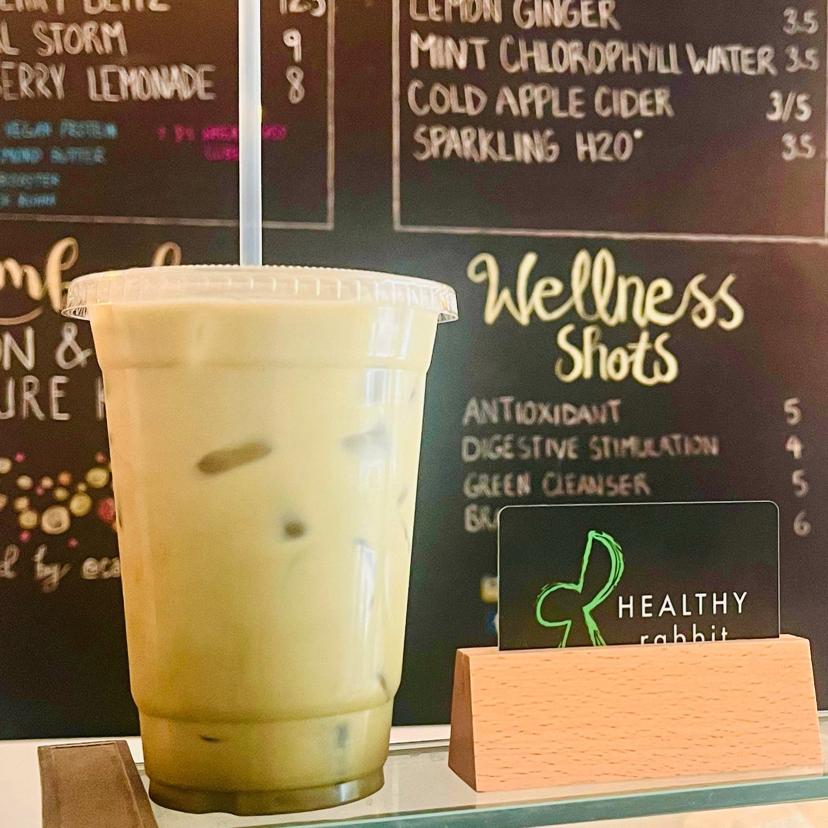 A large plastic cup filled with a green smoothie with ice and a white straw, placed in front of a blackboard menu at a cafe.