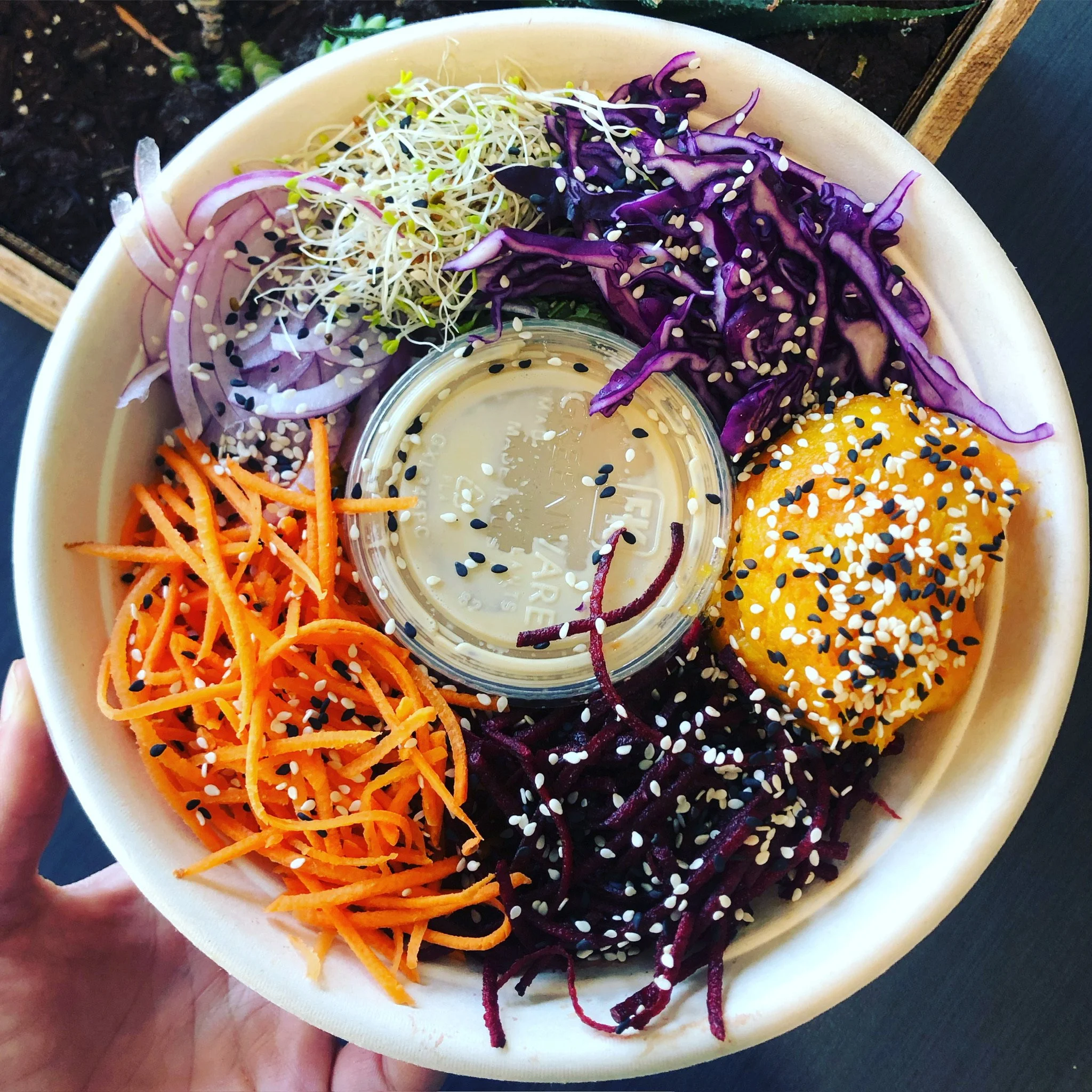 A colorful poke bowl with shredded carrots, purple cabbage, daikon radish sprouts, sliced red onions, and a scoop of spicy mayo topped with sesame seeds, and a small container of dressing in the center.