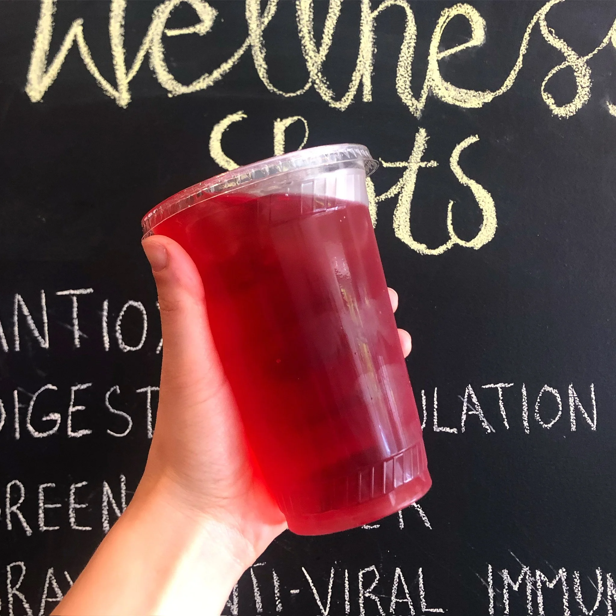 A hand holding a large clear plastic cup filled with red liquid in front of a blackboard with handwritten text about nutrients.