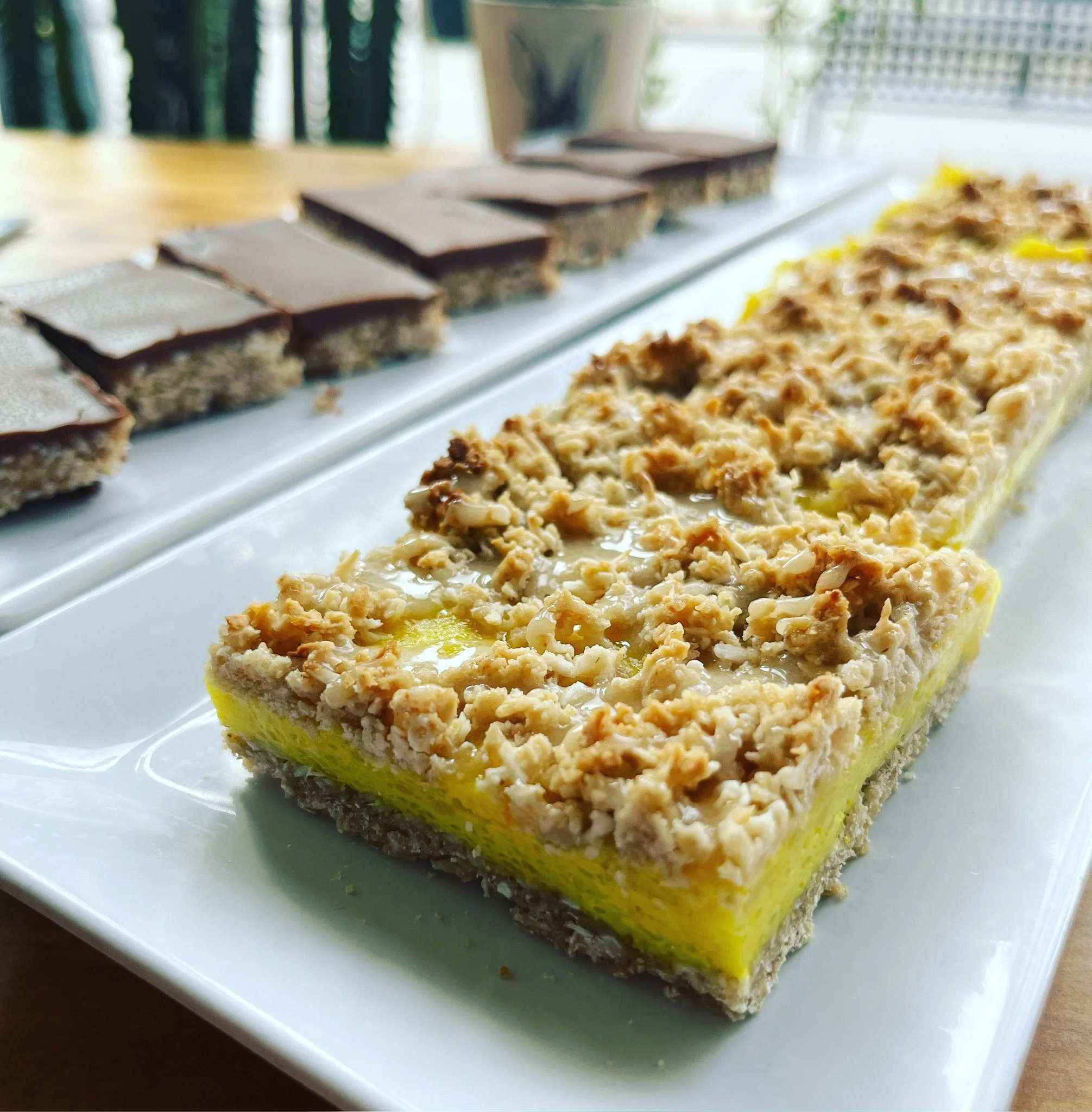 A rectangular dessert bar with a crumbly crust, yellow filling, and a topping of chopped nuts, placed on a white platter. In the background, there are more dessert squares with a layered chocolate topping.