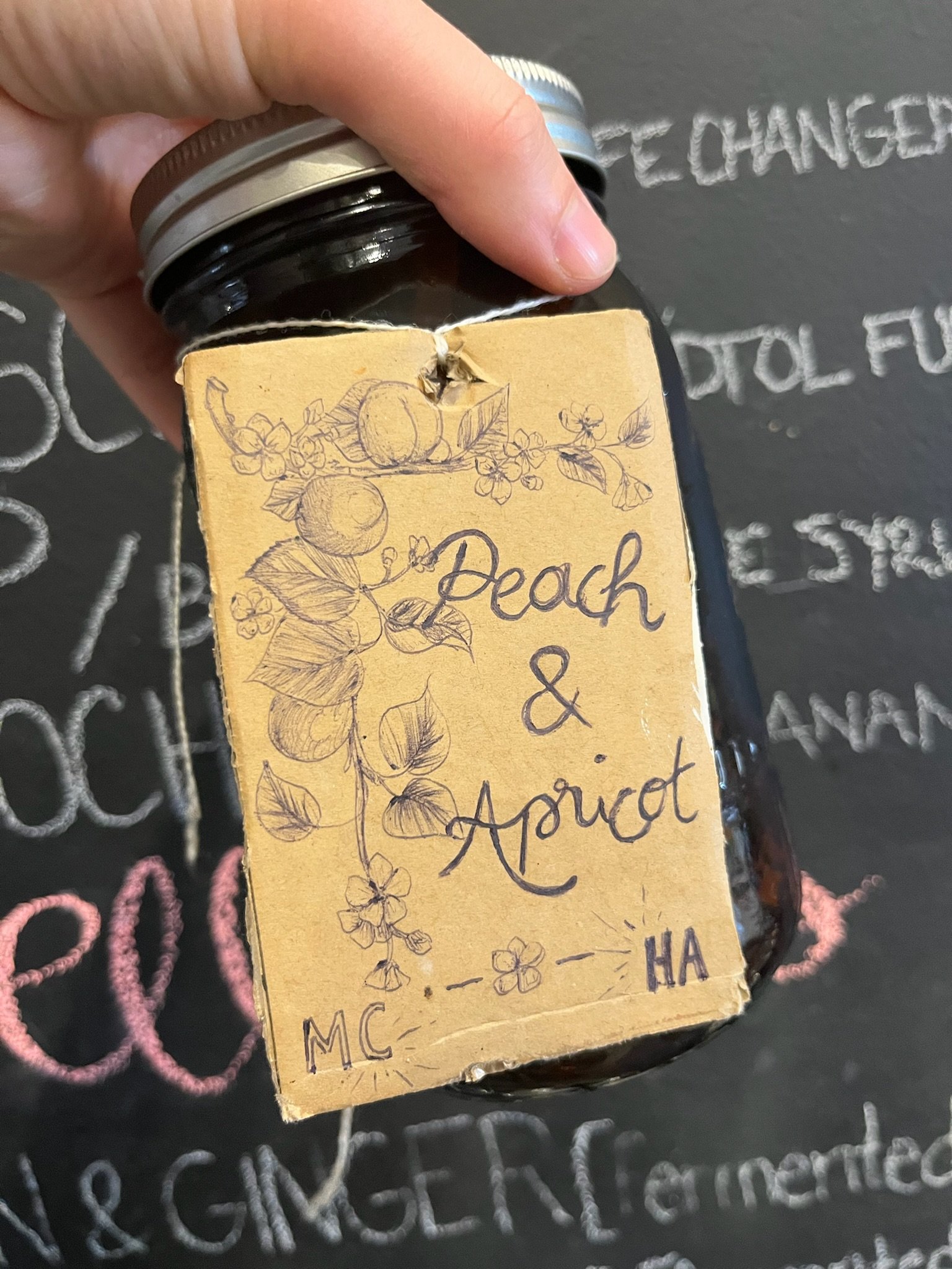 A hand holding a jar of peach and apricot jam with a vintage-style label featuring sketches of peaches, apricots, and flowers, with the handwritten words "Peach & Apricot" and initials "MC HA".