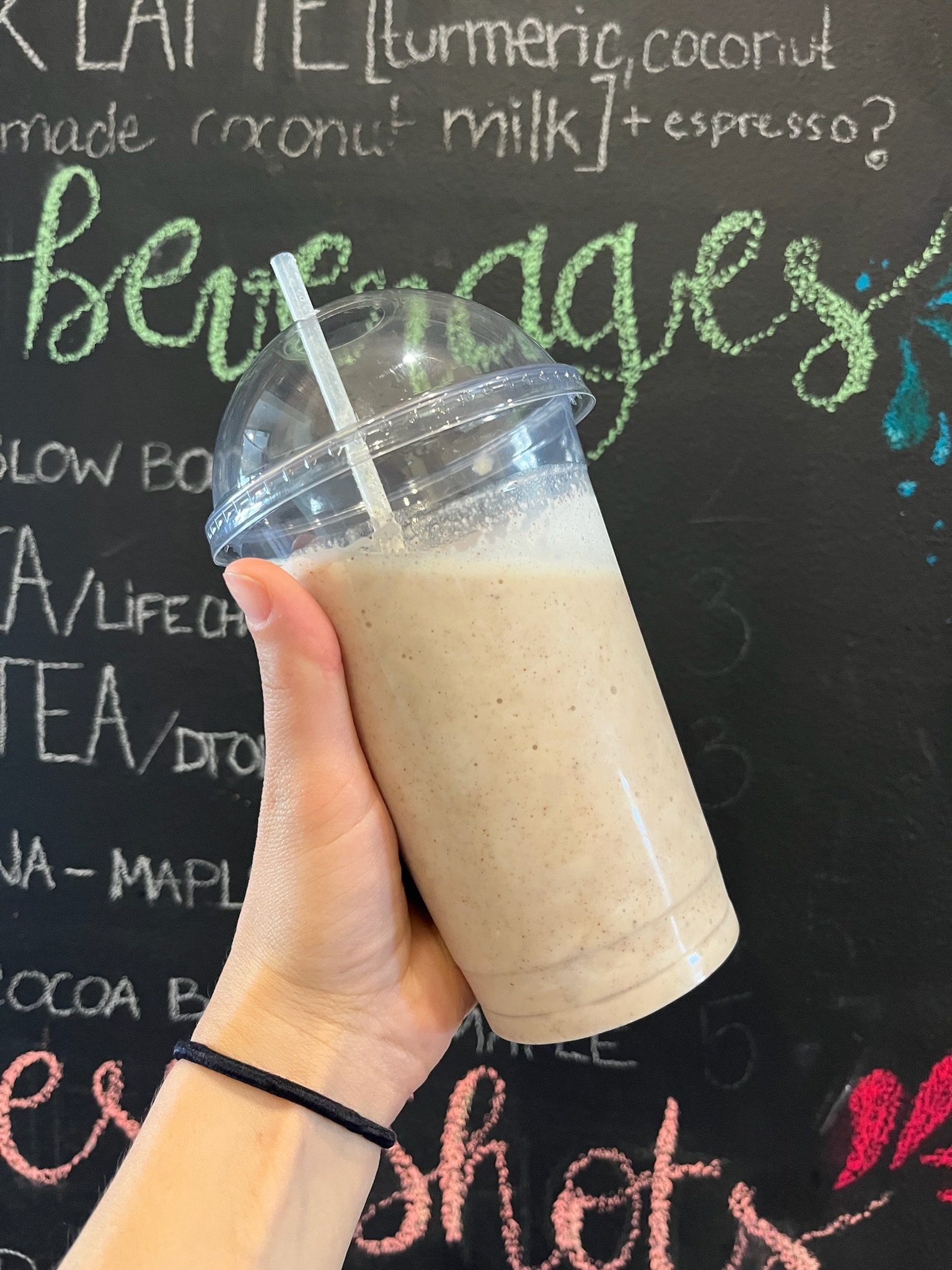 A hand holding a clear plastic cup with a domed lid and straw, filled with a creamy, blended beverage, in front of a black chalkboard with colorful handwritten menu items.