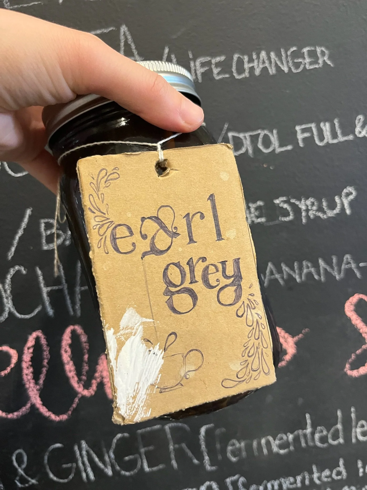 Hand holding a dark glass jar with a beige paper label that says 'earl grey' in handwritten text, attached with a string. In the background, black chalkboard with colorful handwritten recipe ingredients.