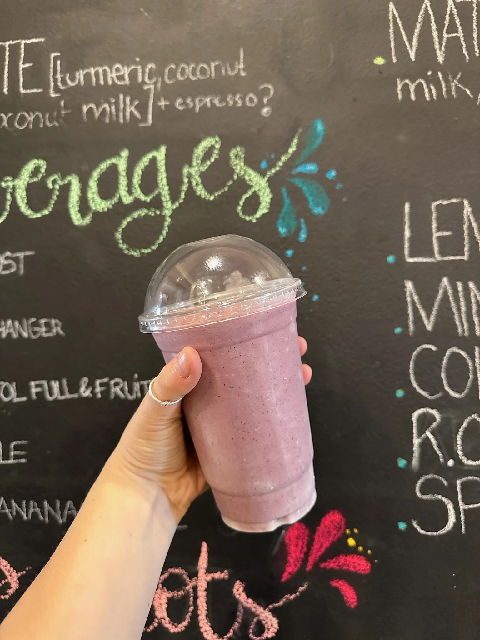 A person holding a pink smoothie in a clear plastic cup with a dome lid in front of a blackboard with colorful chalk writing.