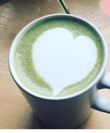 A white mug filled with green latte art in the shape of a heart, on a wooden surface.