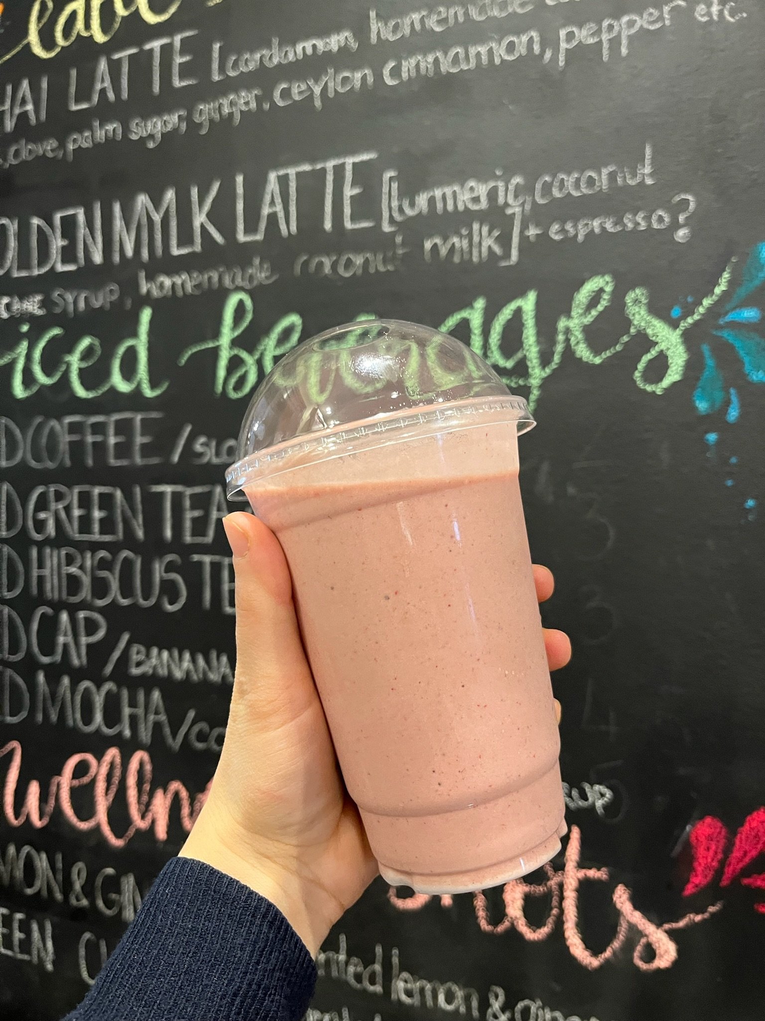 A hand holding a pink smoothie in a clear plastic cup with a dome lid, against a blackboard menu with colorful chalk writing.
