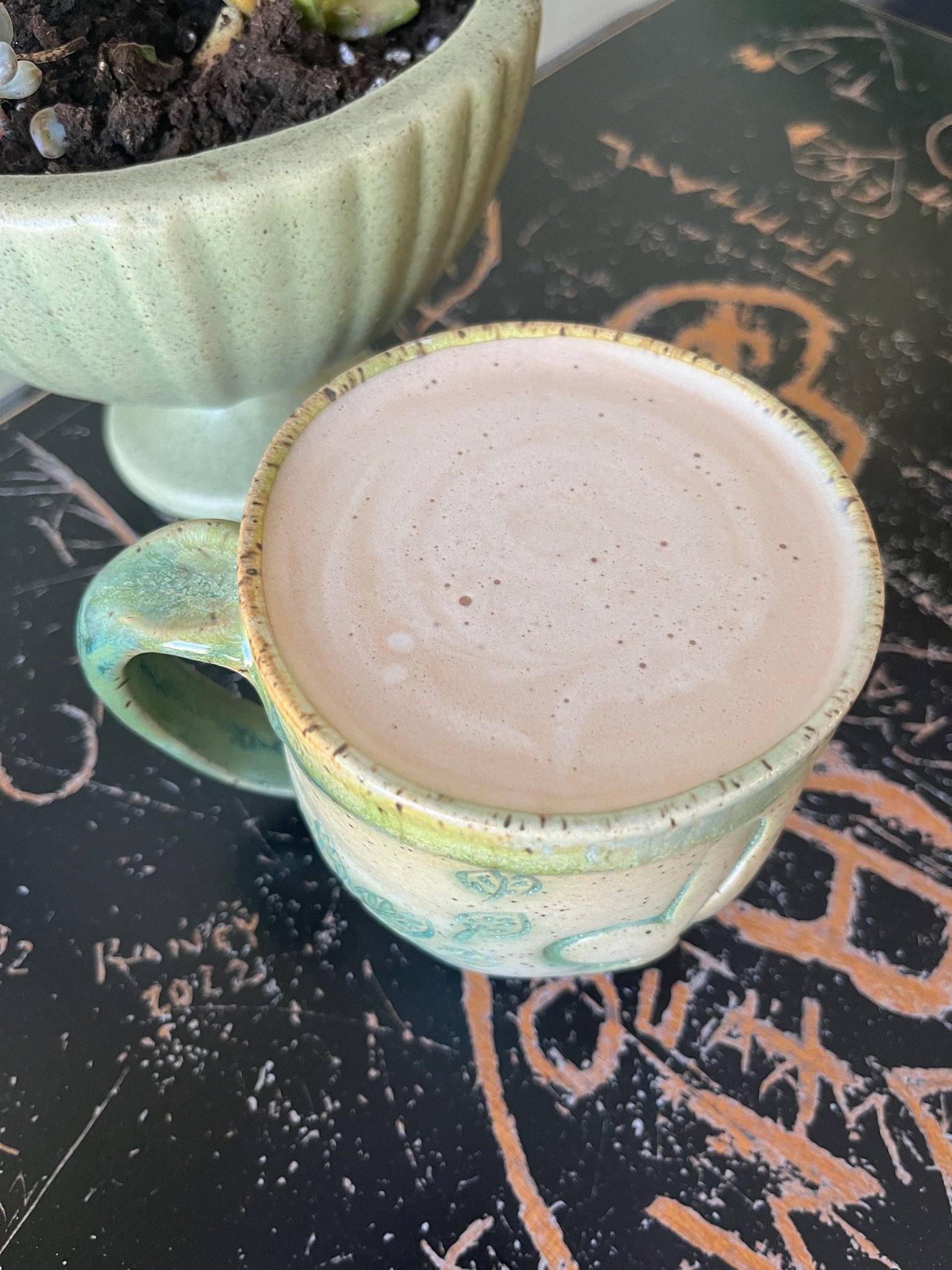 A ceramic mug filled with frothy hot chocolate or coffee with a decorative green and cream pattern, placed on a black chalkboard surface with orange and white chalk writing and a potted succulent plant in a green ceramic planter in the background.