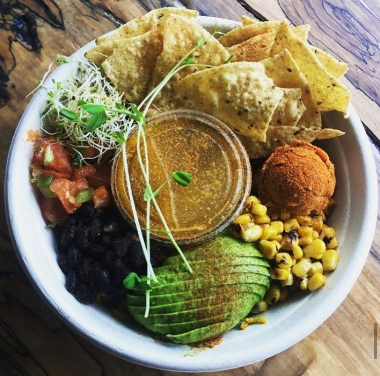 A bowl of Mexican food with tortillas, avocado slices, corn, beans, pico de gallo, microgreens, a ball of fried cheese, and a small cup of sauce.