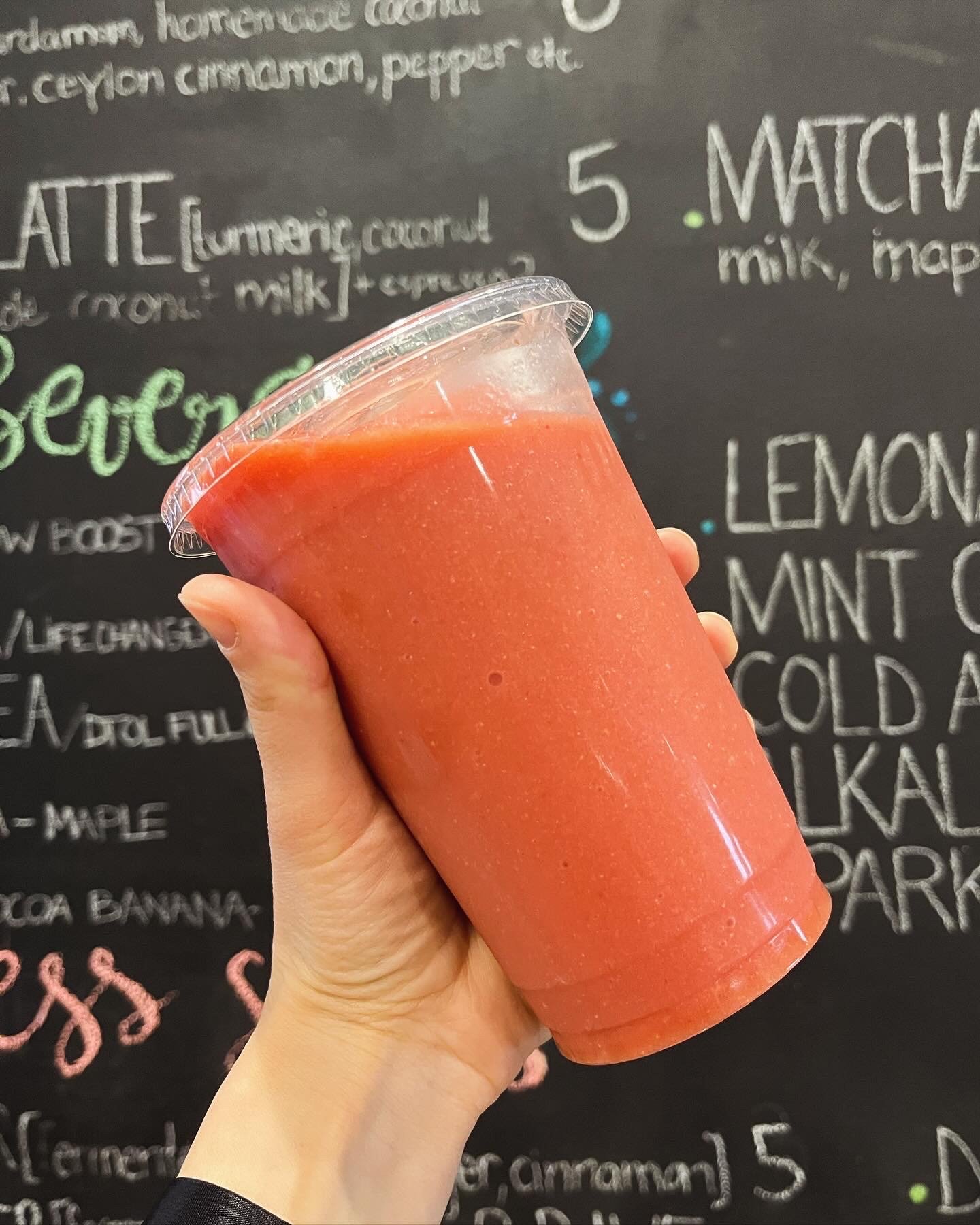 A hand holding a red smoothie in a clear plastic cup in front of a black chalkboard menu with white and colorful writing.