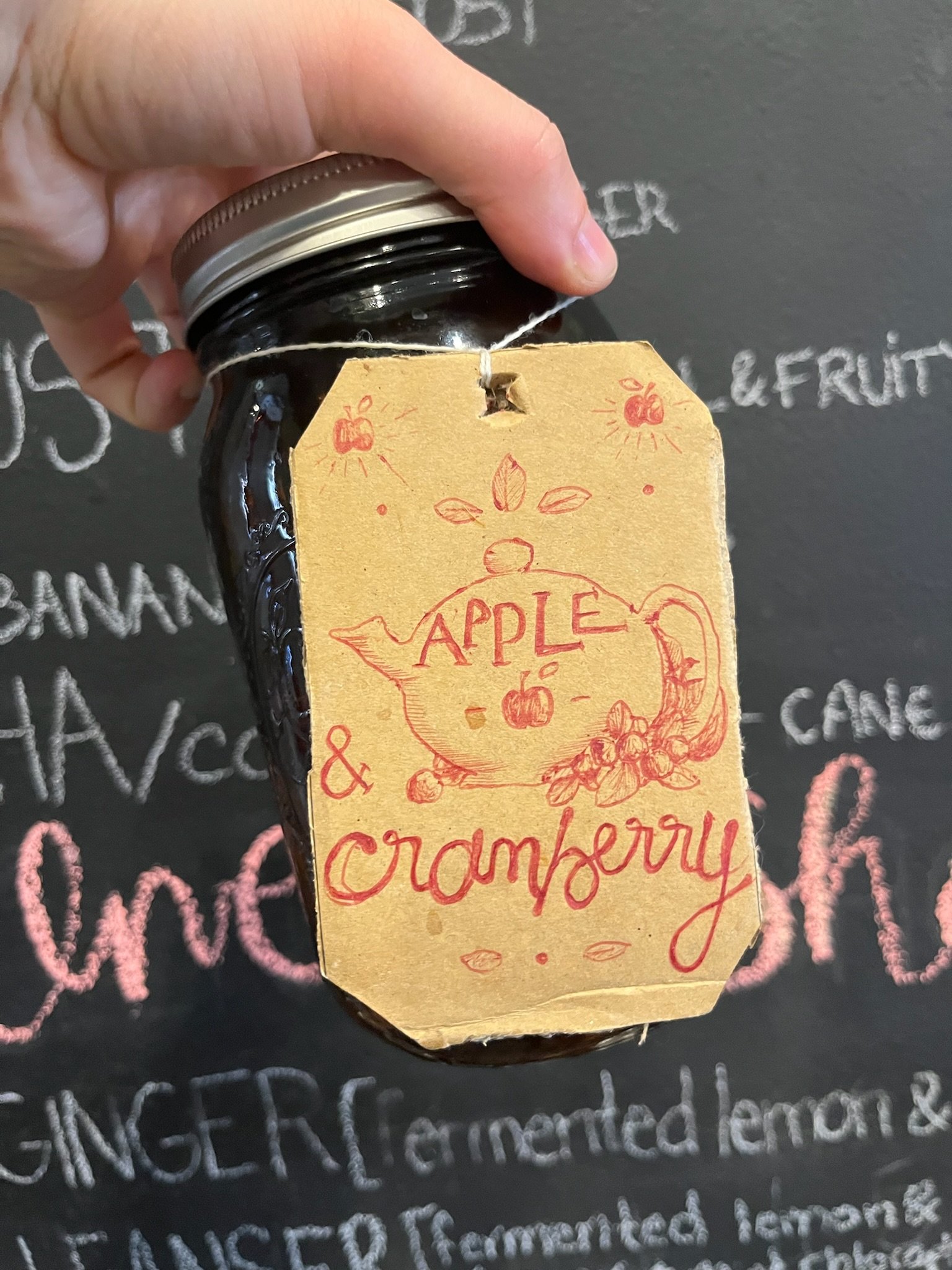 Close-up of a hand holding a jar of apple and cranberry jam, with a decorative beige tag attached by a string, featuring a red illustration of a teapot and handwritten-style text reading 'Apple & Cranberry'.