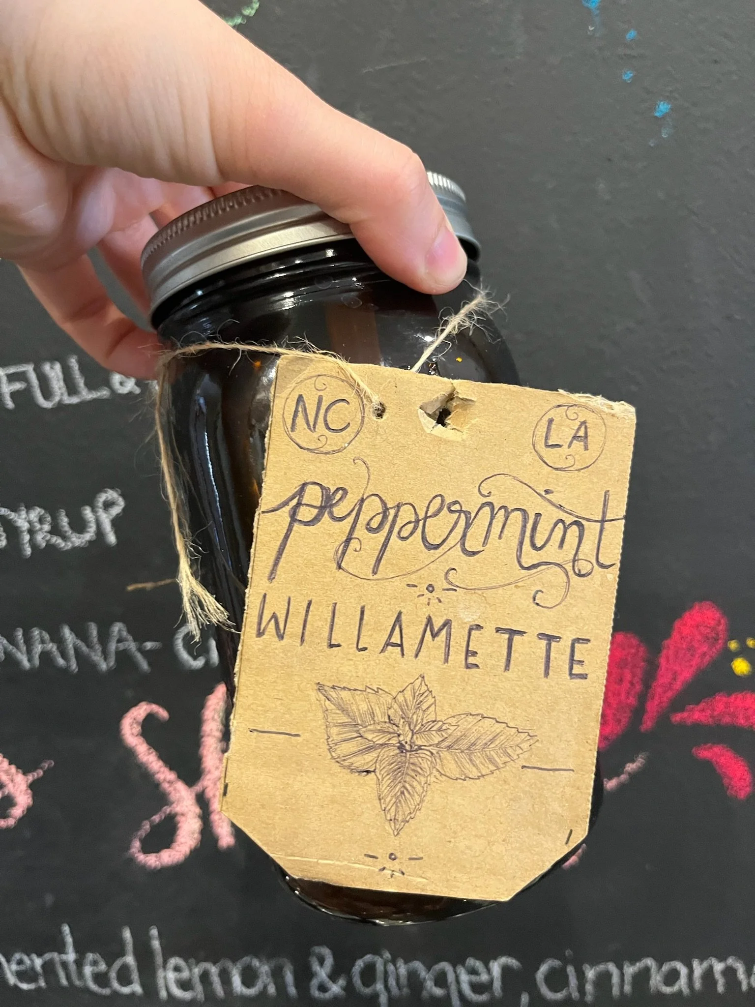 A person's hand holding a glass jar with a handwritten label that says 'North Carolina' and 'Willamette.' The label also features a drawing of a mint leaf and decorative handwriting.