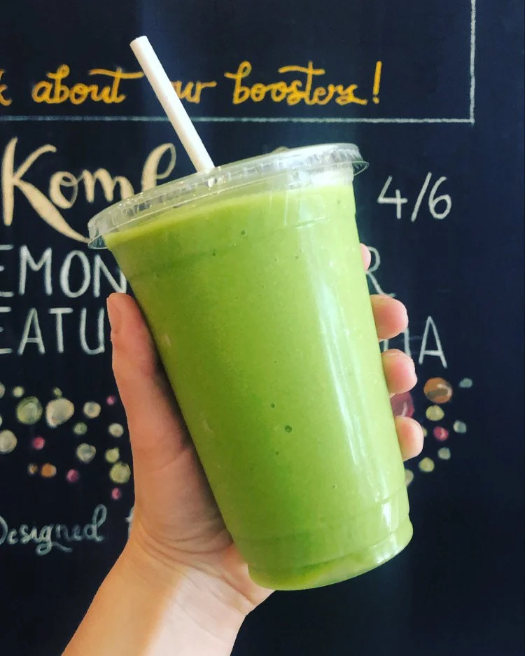 A hand holding a clear plastic cup filled with a bright green smoothie, with a white straw, against a blackboard background with colorful writing and drawings.