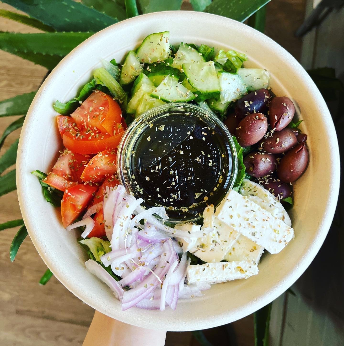 A bowl of Greek salad with cucumbers, tomatoes, Kalamata olives, red onions, feta cheese, and a small container of dark salad dressing or balsamic vinegar, topped with herbs.