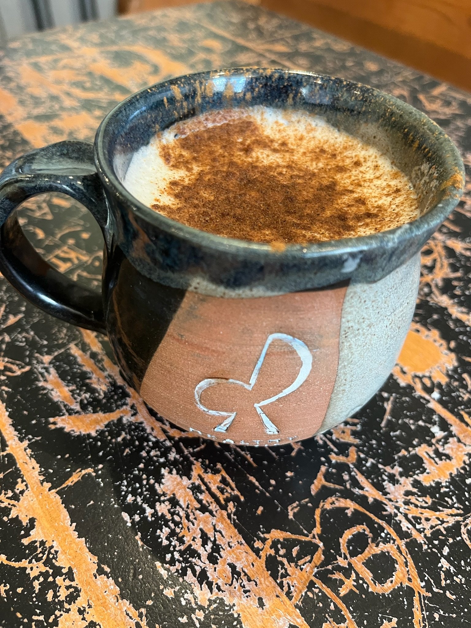A ceramic mug filled with a hot beverage topped with cinnamon or cocoa powder, resting on a black and orange speckled table.