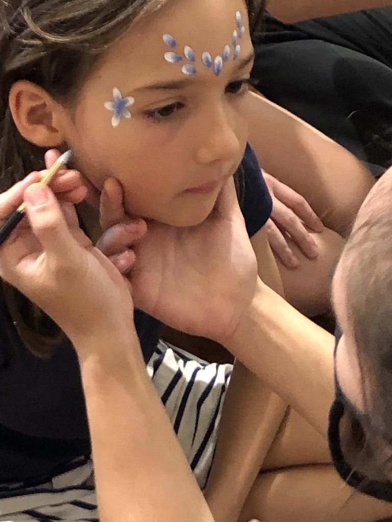 A young girl having her face painted with blue and white flower designs by an artist, with the artist's hands visible holding a paintbrush.