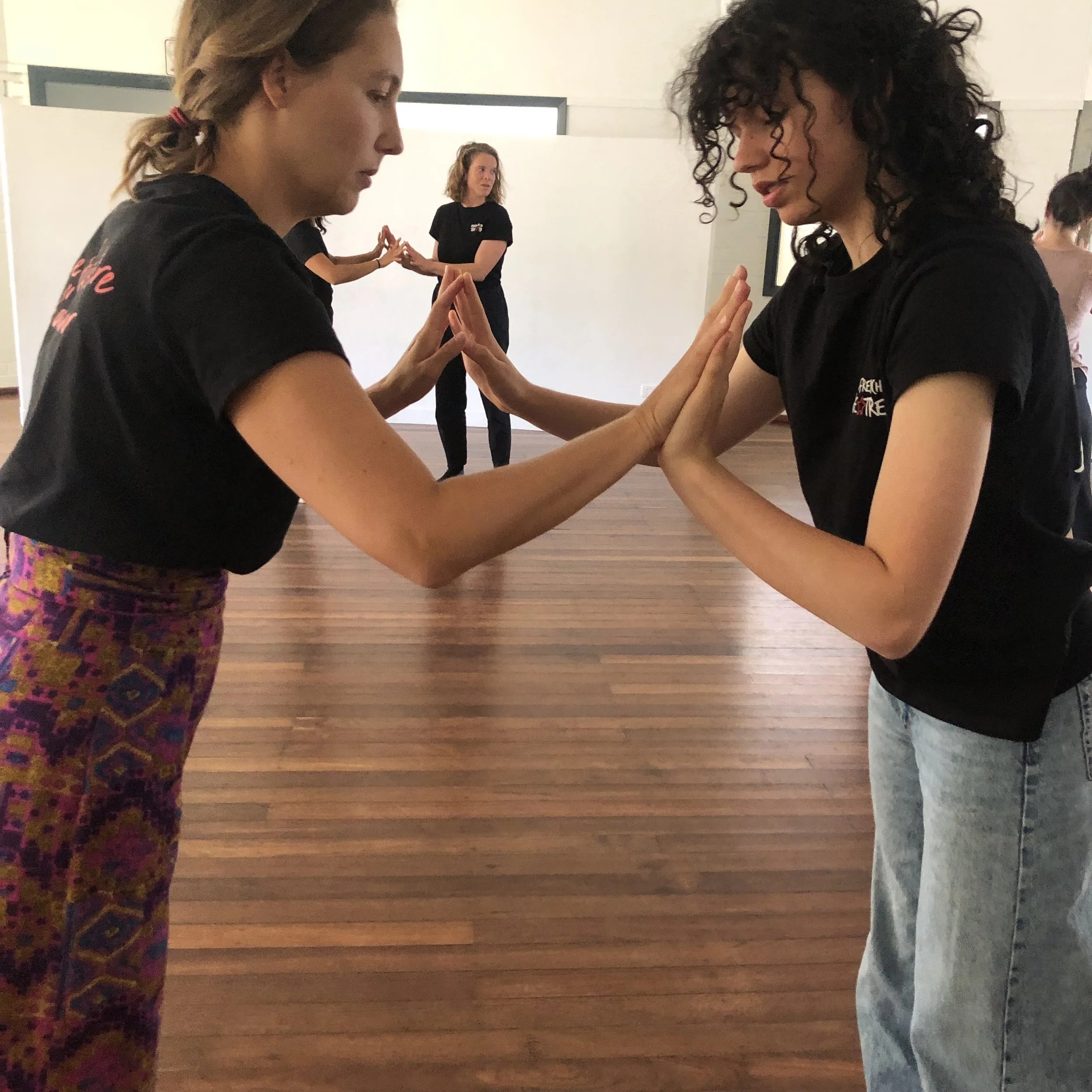 Two participants facing each other during a Perth French Theatre movement exercise, with others rehearsing in the background.