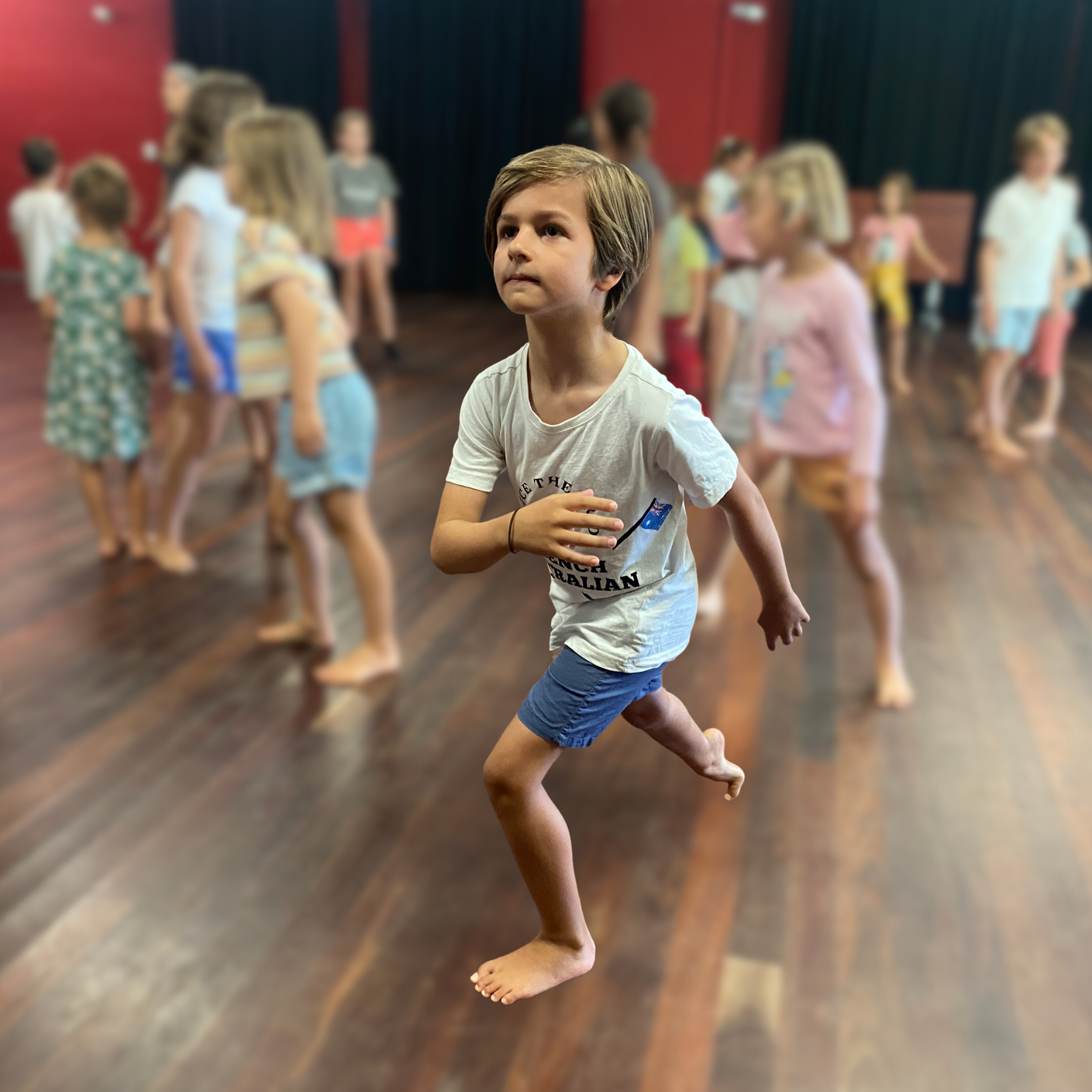 A young boy with brown hair, wearing a white T-shirt and blue shorts, is jumping in a room with a group of children in the background. The children are barefoot and appear to be playing or dancing.