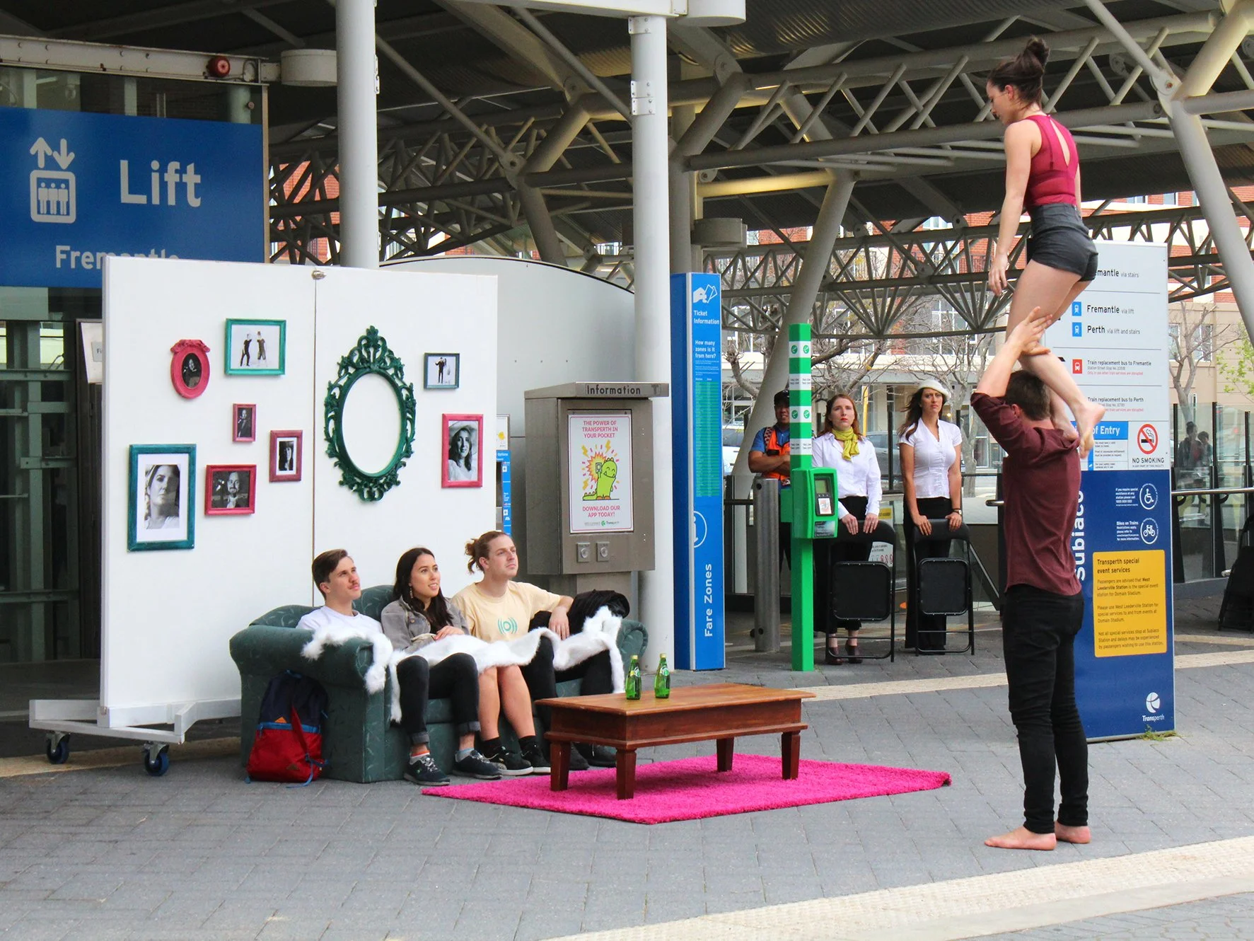 A group of four young adults sitting on a dark green couch at a train station, watching a performer. The performer, barefoot, is balancing on one foot on a male's hand, which is raised above his head, as part of a fire or street act. There is a pink rug under the performer and a small wooden table with two green bottles nearby. In the background, there are people standing and waiting, with signs indicating train directions, and decorations on a white panel.