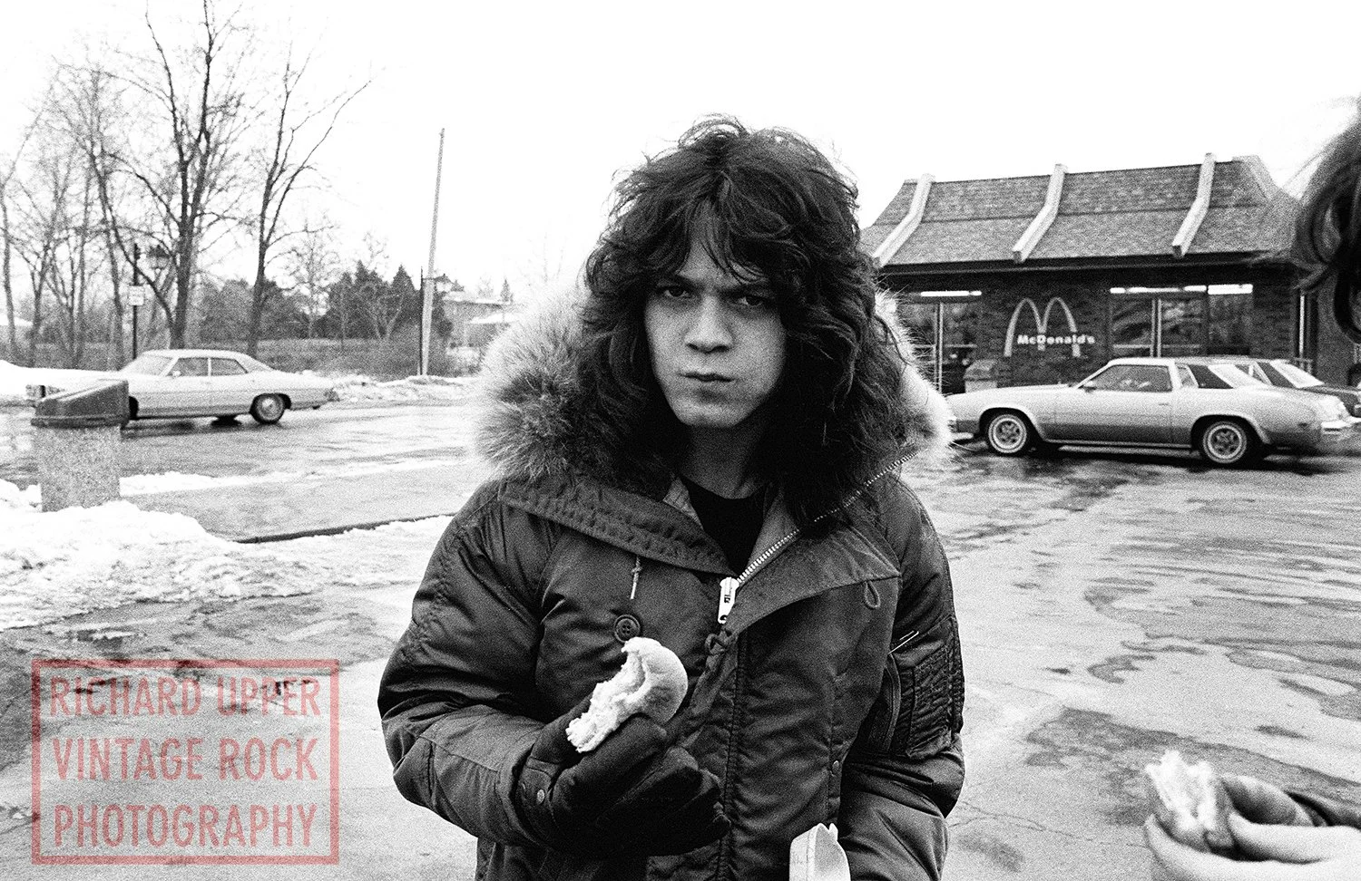 Black and white photo of a young person with curly hair wearing a winter jacket with a fur-lined hood, standing outdoors in a parking lot with snow and ice. A McDonald's restaurant is visible in the background.
