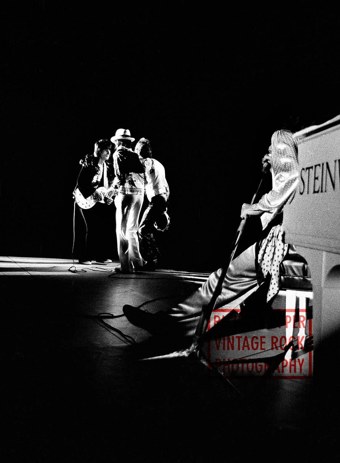Black and white photo of a band performing on stage with four members, one playing guitar, one singing into a microphone, and others standing close together. A person is seated near a vintage Steingarten piano, partially visible on the right.