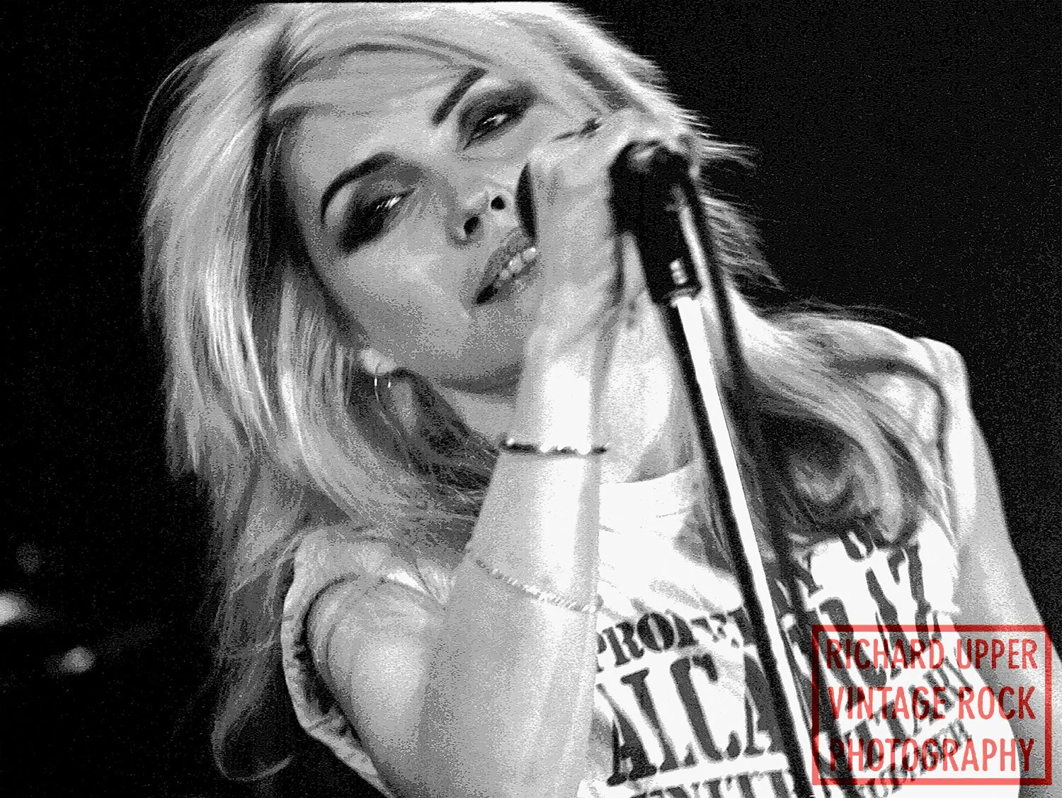 Black and white photo of a female singer with long wavy hair singing into a microphone, wearing a sleeveless top with text, and a bracelet on her wrist.
