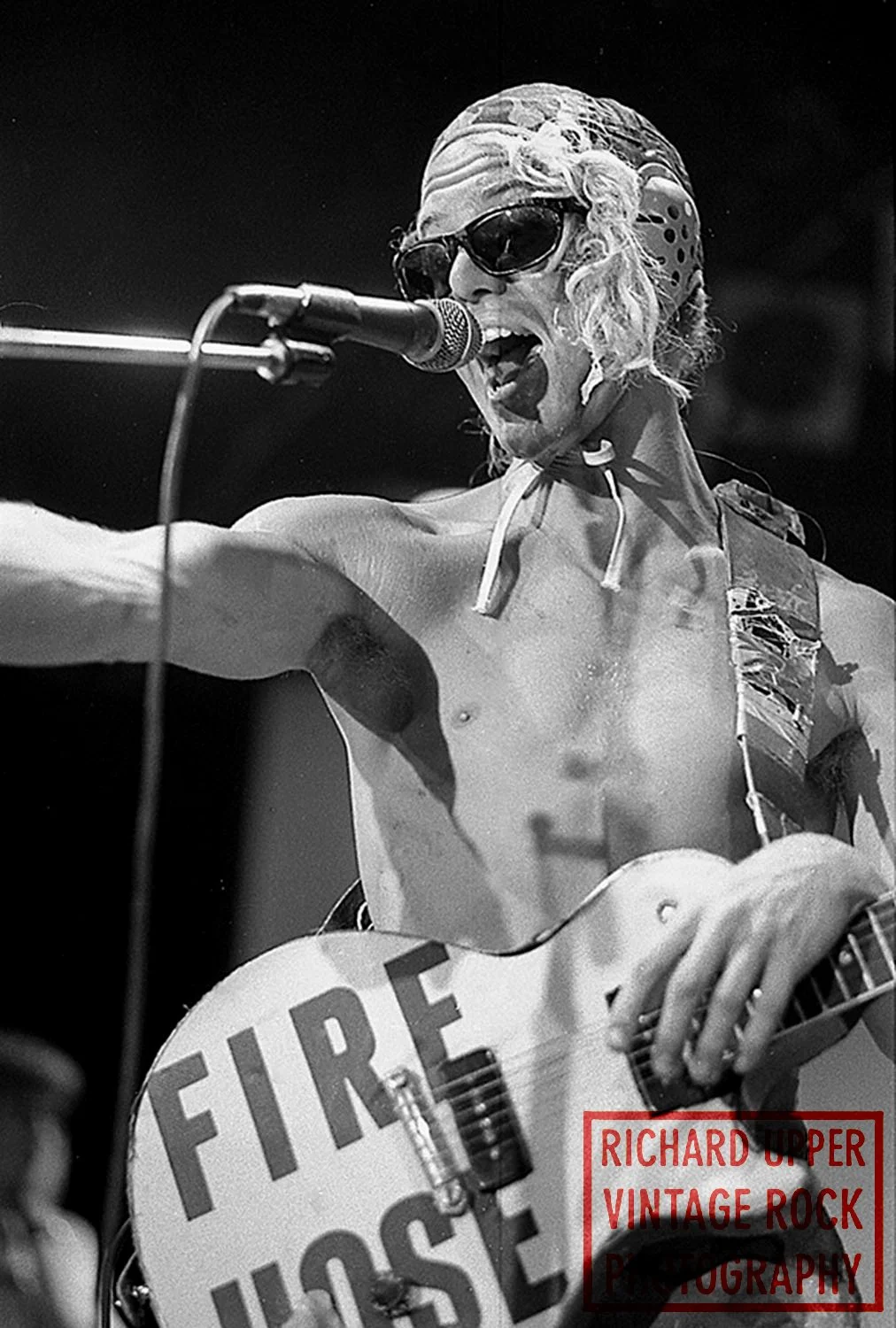 Black and white photo of a shirtless musician with long hair, sunglasses, and headphones, singing into a microphone while playing an electric guitar with a sign that says 'FIRE' on it, with text 'RICHARD UPPER VINTAGE ROCK PHOTOGRAPHY' in the corner.