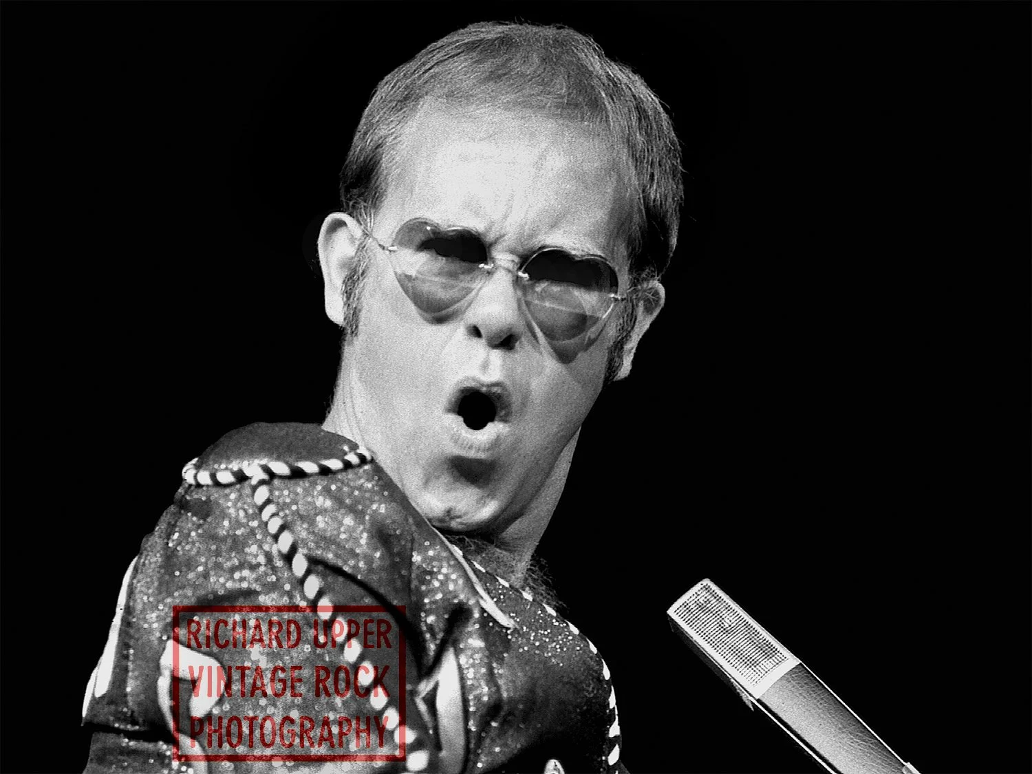 Black and white photo of a man with short hair wearing sunglasses and a patterned jacket, singing into a microphone with an expression of surprise or excitement.