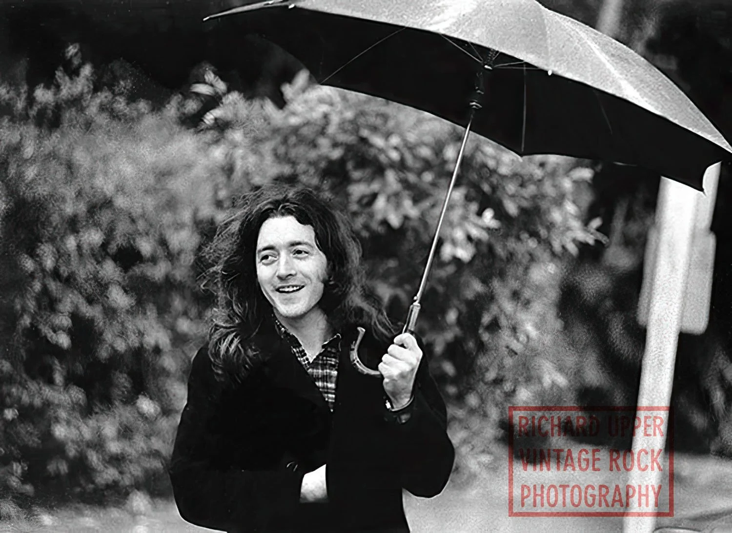 A black and white photograph of a man with long, wavy hair holding an umbrella outdoors. He's wearing a checkered shirt and a dark jacket, smiling and looking to the side with a background of blurred foliage. The image is credited to Richard Upper Vi
