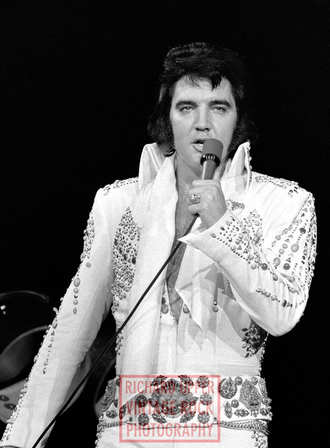 Black and white photo of Elvis Presley holding a microphone and singing, wearing a sequined white jumpsuit with decorative embellishments.