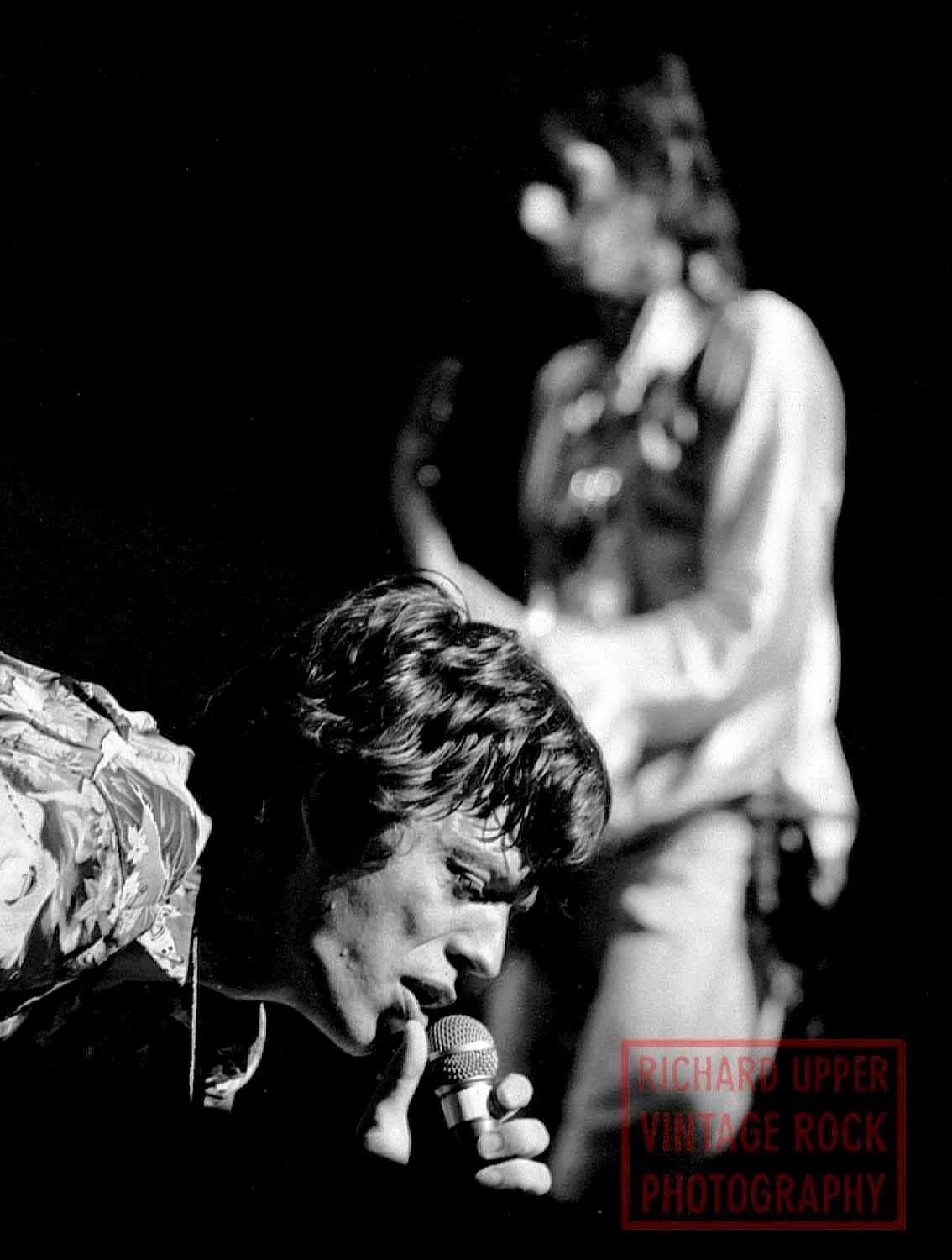Black and white photo of a woman singing into a microphone, with two men in the background. The photo is credited to Richard Upper Vintage Rock Photography.