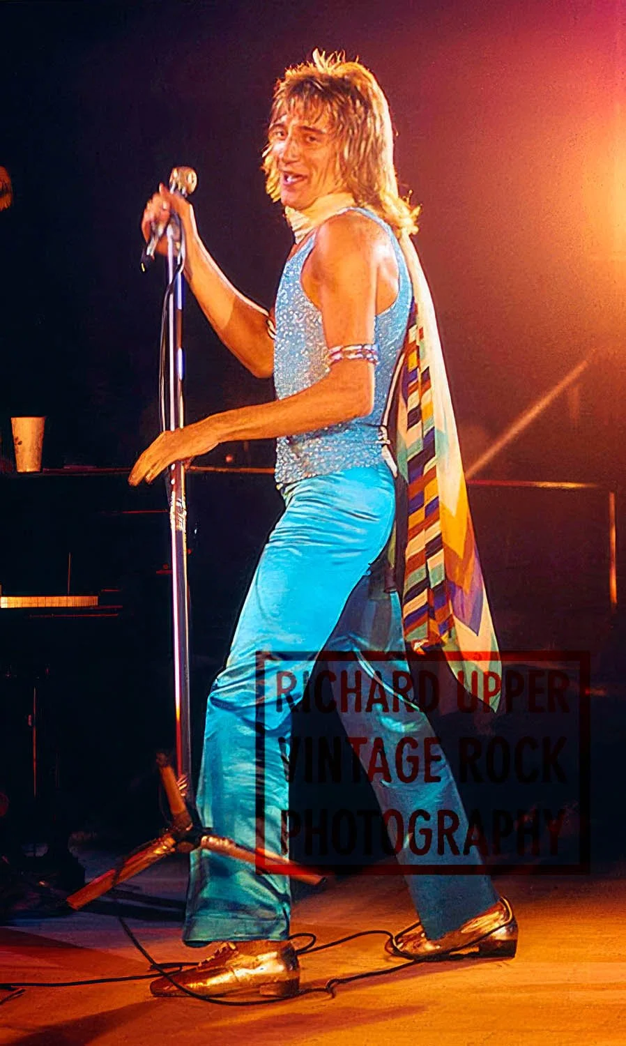 Male performer singing on stage with a microphone, wearing shiny blue pants, a sparkly tank top, metallic shoes, and a colorful scarf draped over his shoulder, with stage lighting in the background.