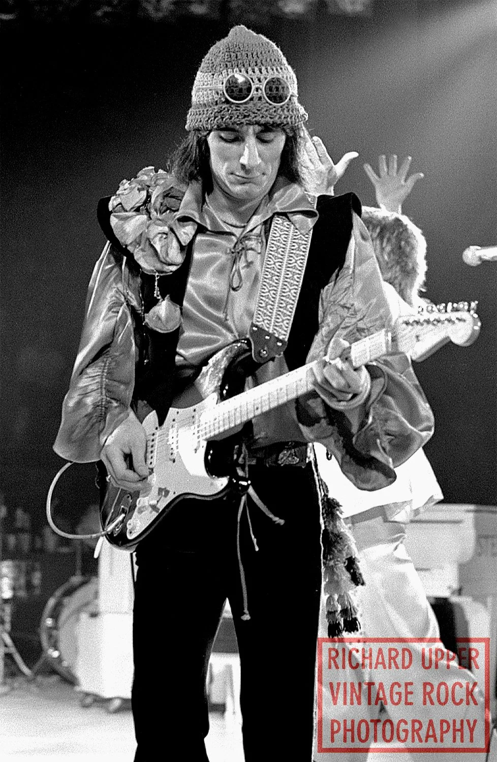 A black and white photo of a person wearing a knit hat with sunglasses, playing an electric guitar, with other musicians in the background. The image has a watermark that reads "RICHARD UPPER VINTAGE ROCK PHOTOGRAPHY."